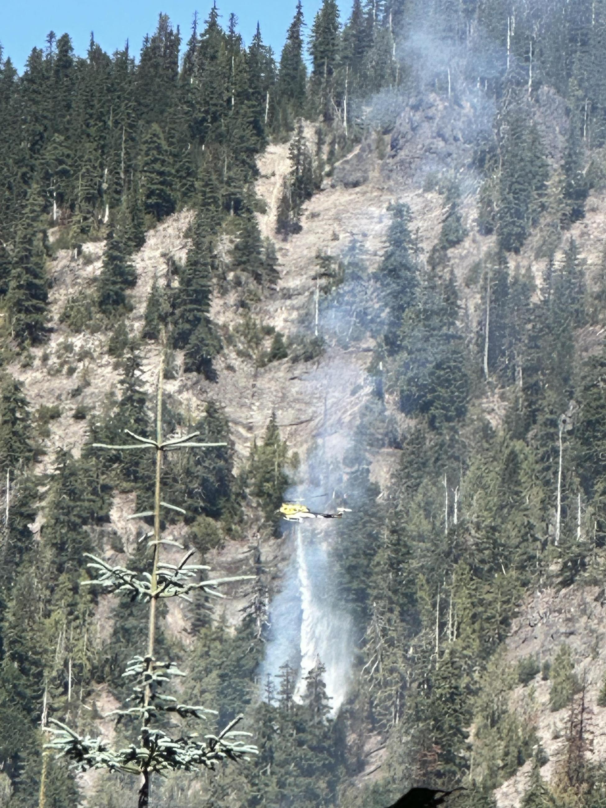 Water drops onto burning vegetation against a hillside of rocks and trees.