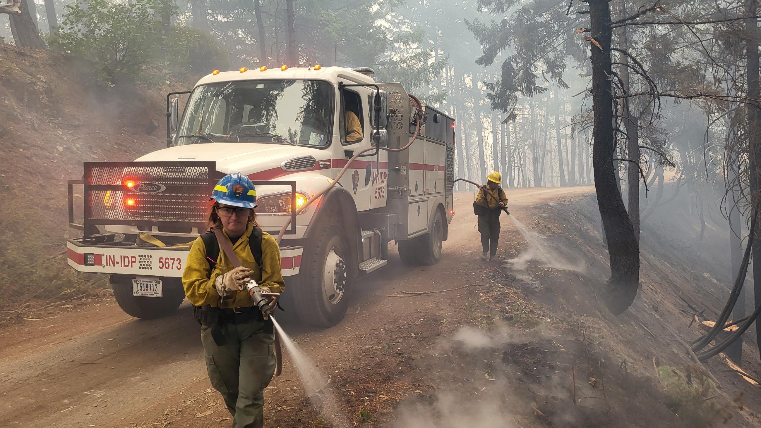 A type 3 red and white fire truck with two firefighters dressed in Nomex wet mop along the road side after a back burn. 