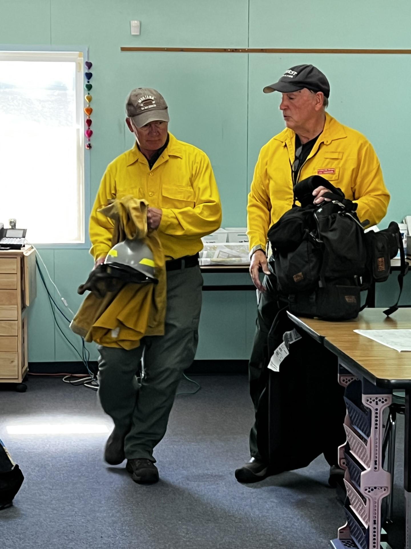Two PIOs hold fireline equipment in a classroom