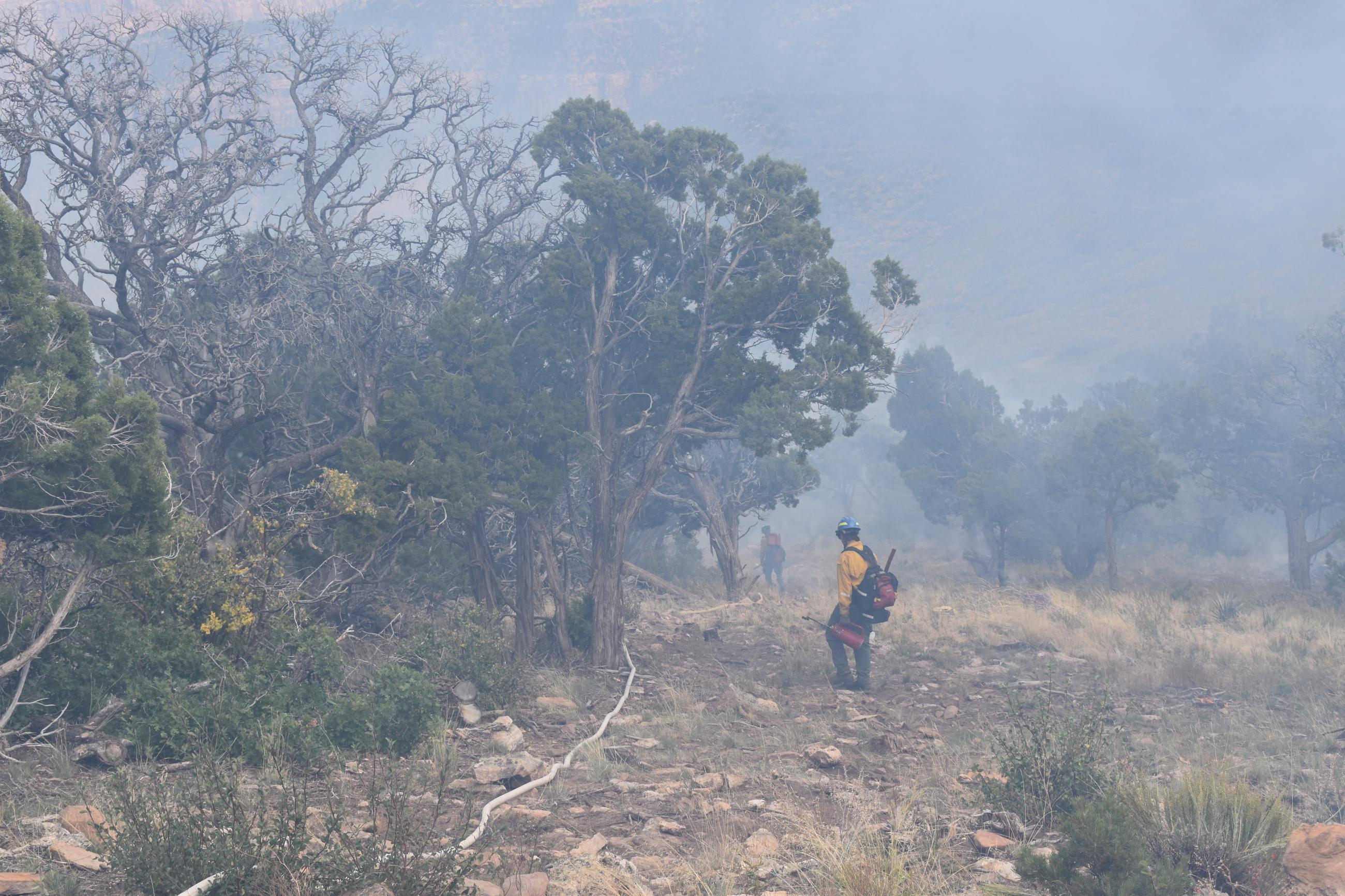 Image of two firefighters securing the north line with hose lay, juniper and pinyon pine trees, rocks, grass, and smoke.