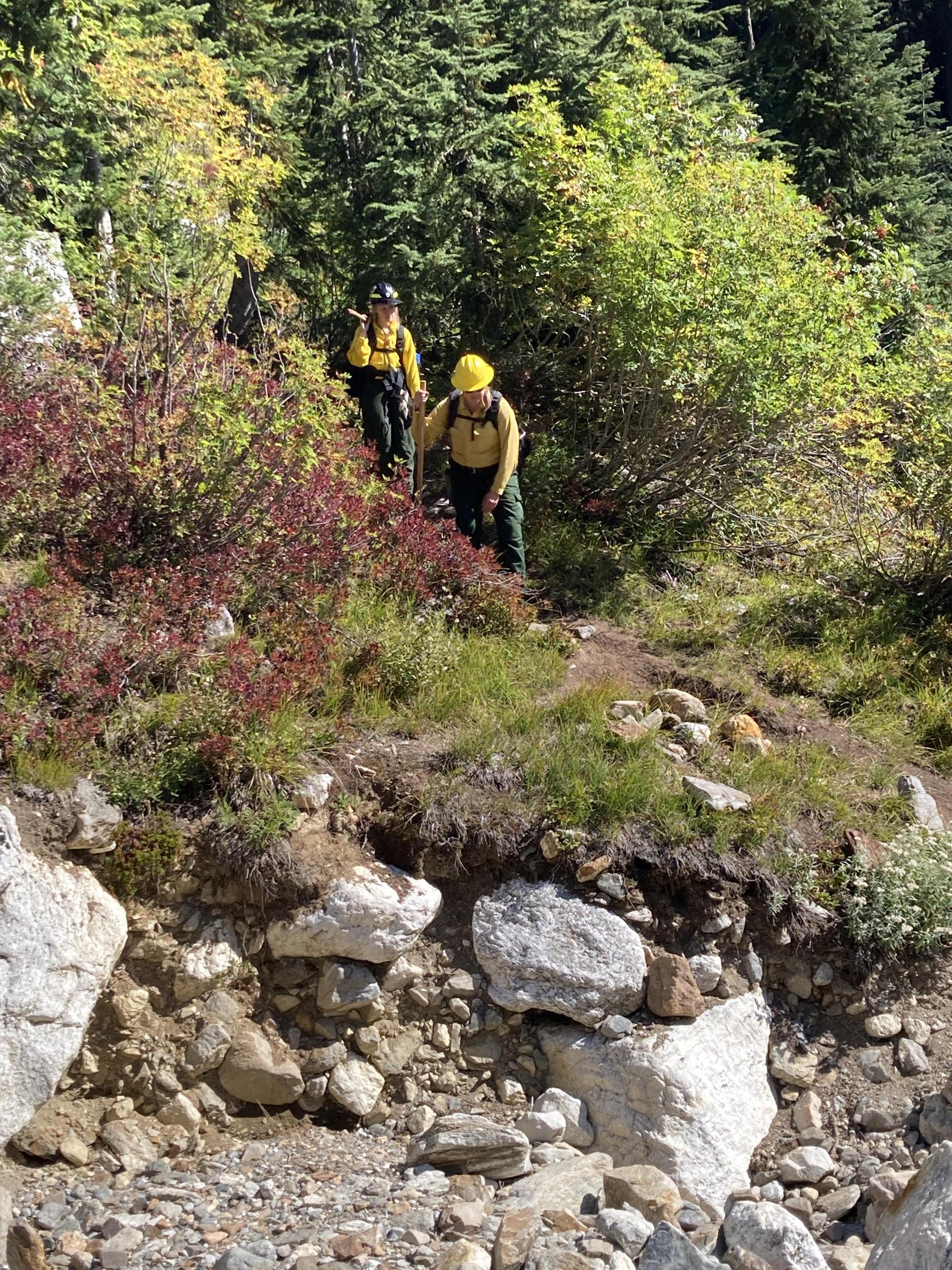 Two women in yellow shirts, green pants, and helmets hike through dense shrubs in rocky terrain