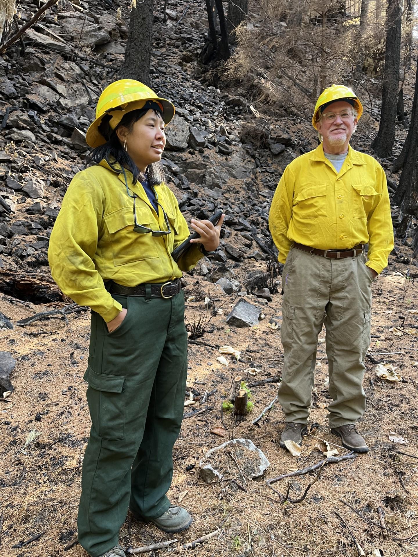 A woman in yellow helmet, yellow shirt, and green pants stands beside a man in yellow shirt, yellow helmet, and tan pants on bare soil with burnt trees behind them