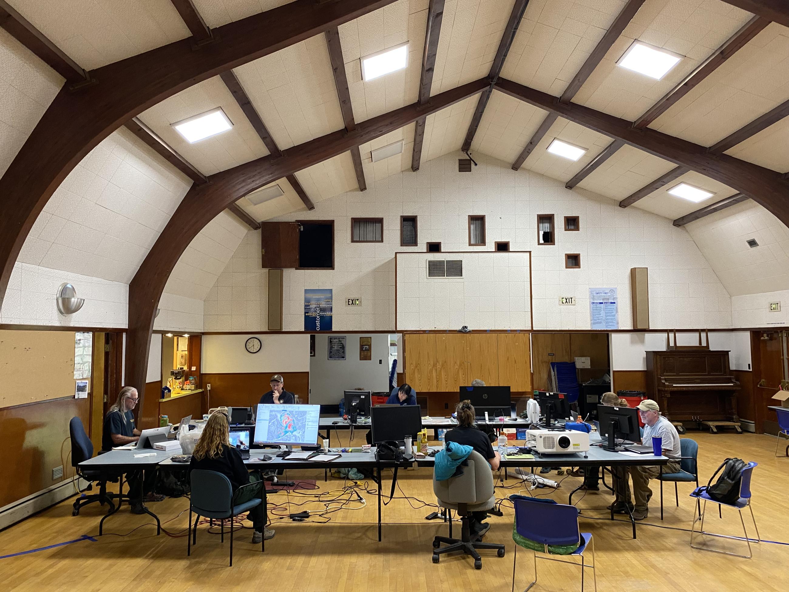 Several people work at computers placed at tables in a big room with artificial lighting
