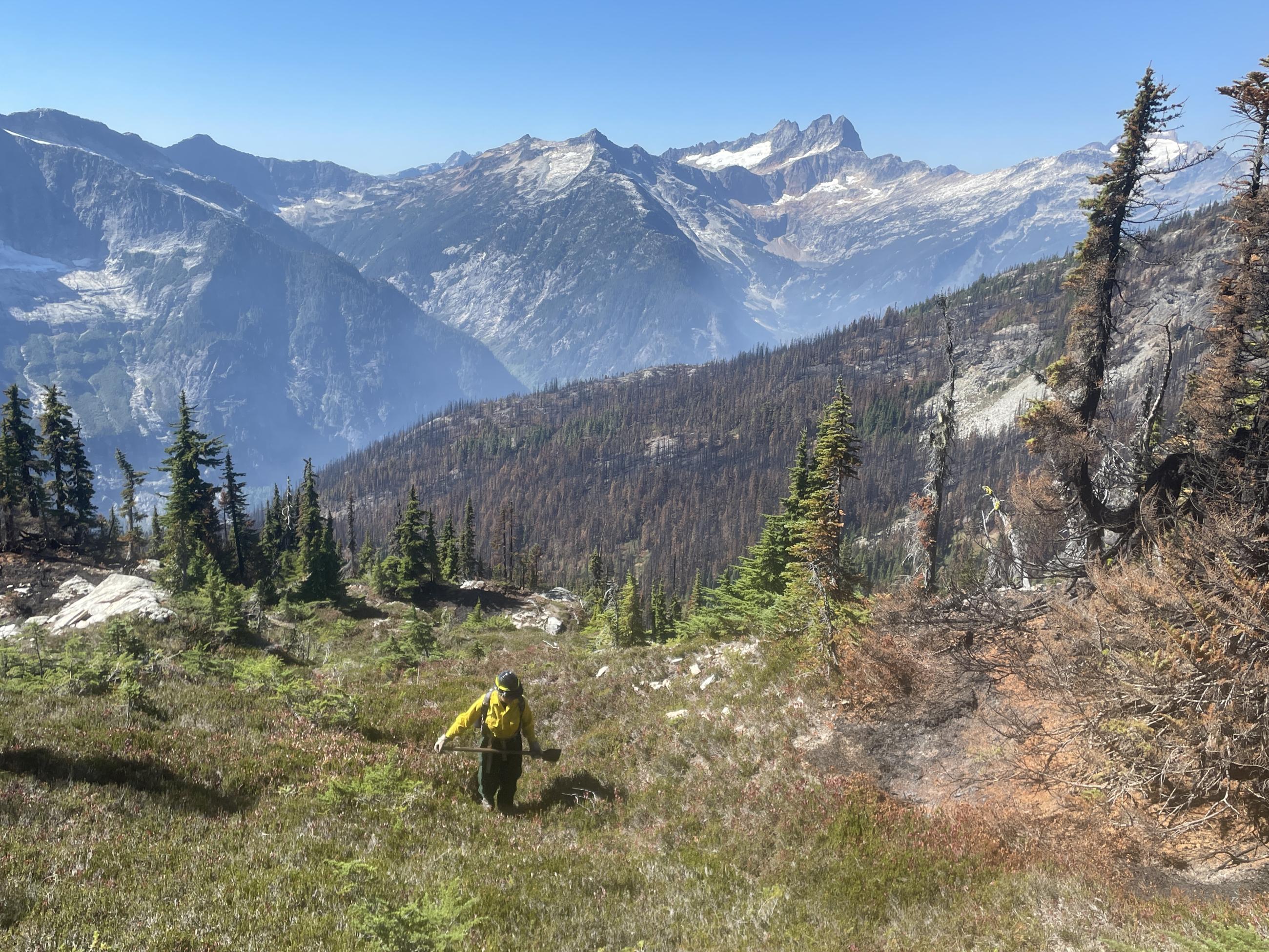 A scientist in firefighting gear holds a shovel and walks through a rocky area in front of mountains