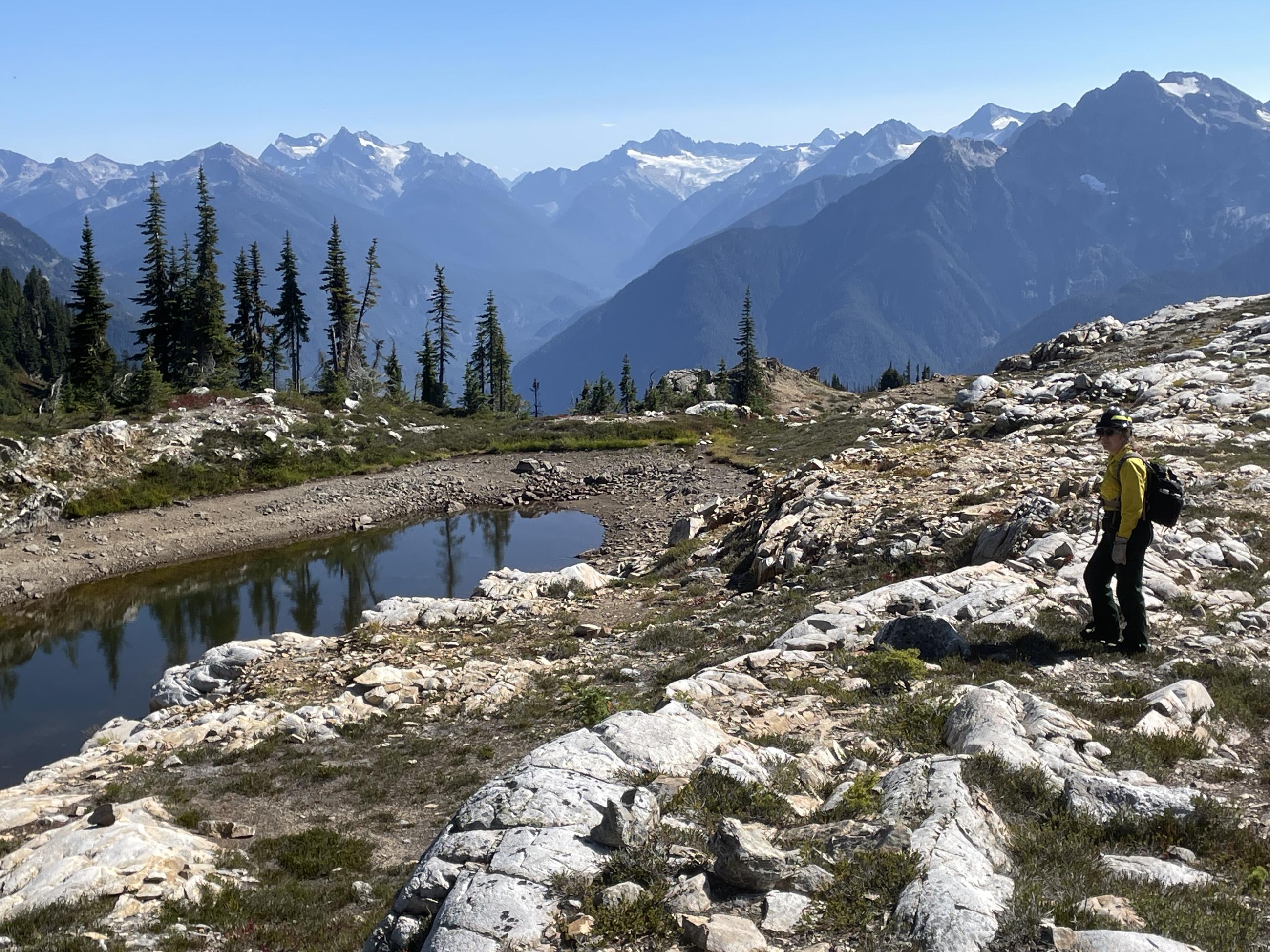 A woman wearing a yellow shirt, green pants, and helmet stands with a hiking stick on rocky terrain with mountains in the background