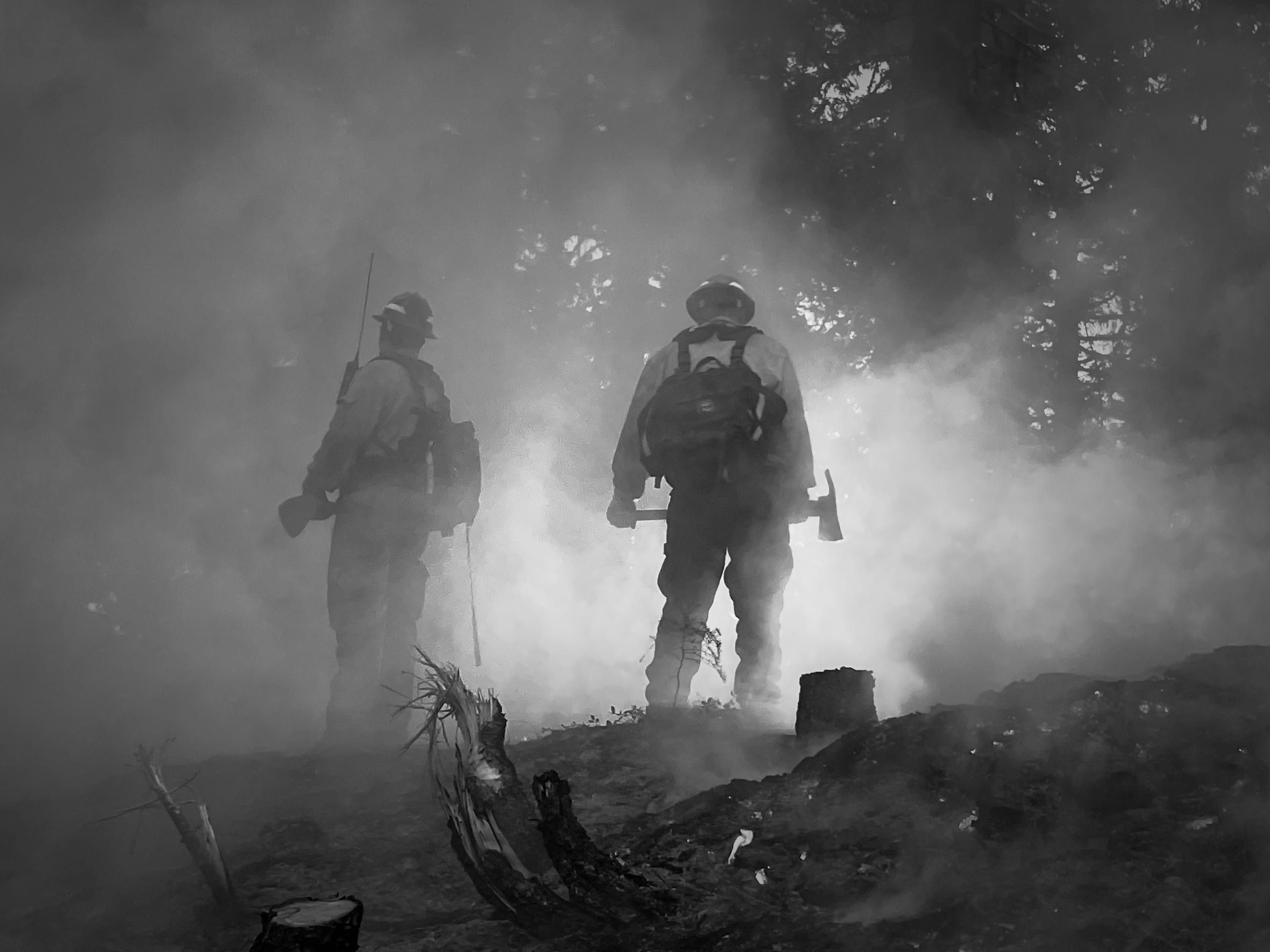 a black and white photo of two firefighters obscured by smoke working with handtools