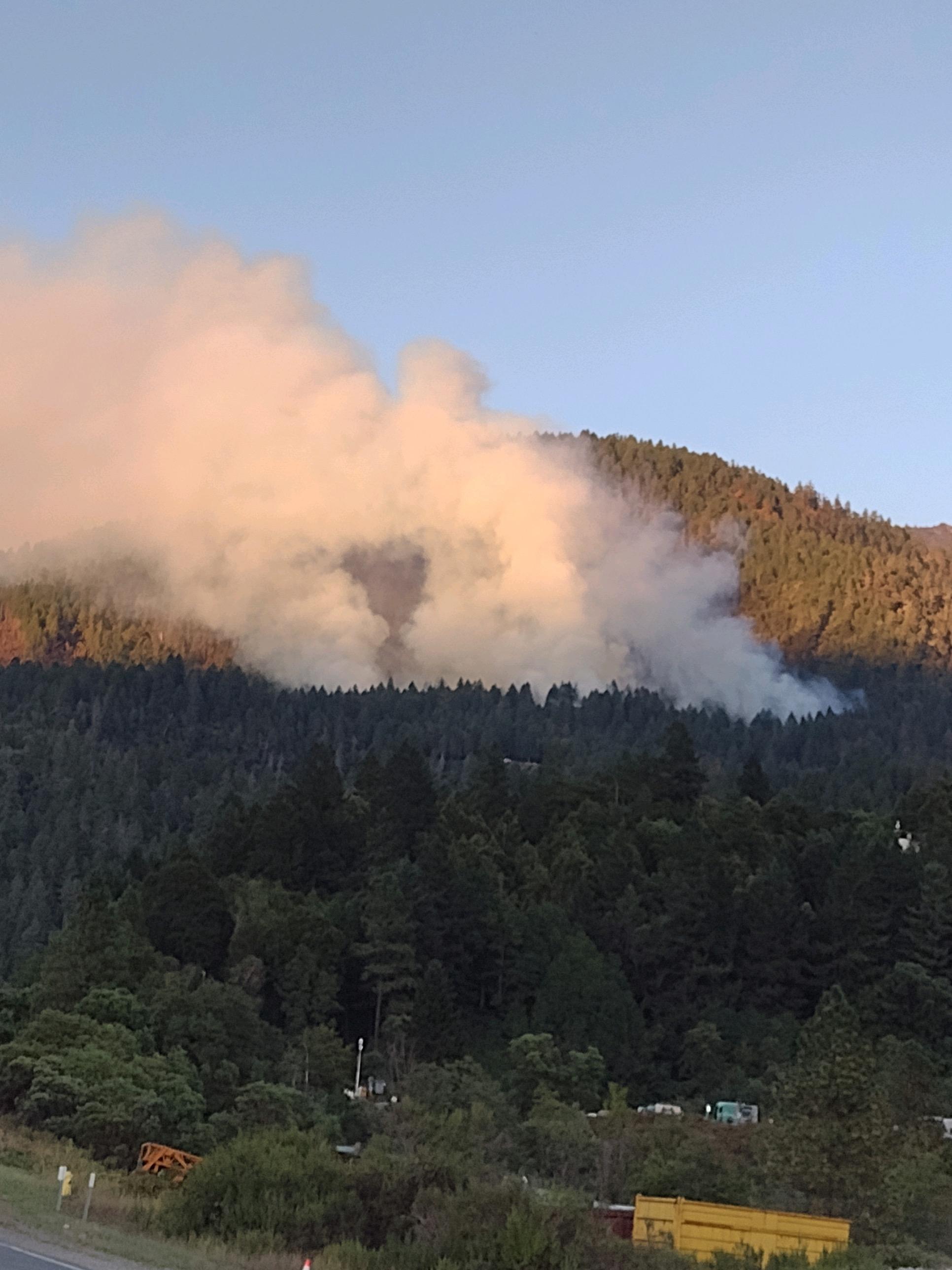 Large white smoke plume blowing East between two forested mountains