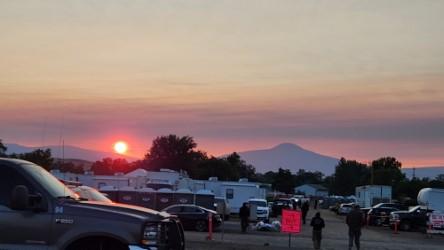Image of the sun rising behind the fire camp with tents, trailers, vehicles and people walking in the camp. Photo USDA Forest Service courtesy Donavun Davis