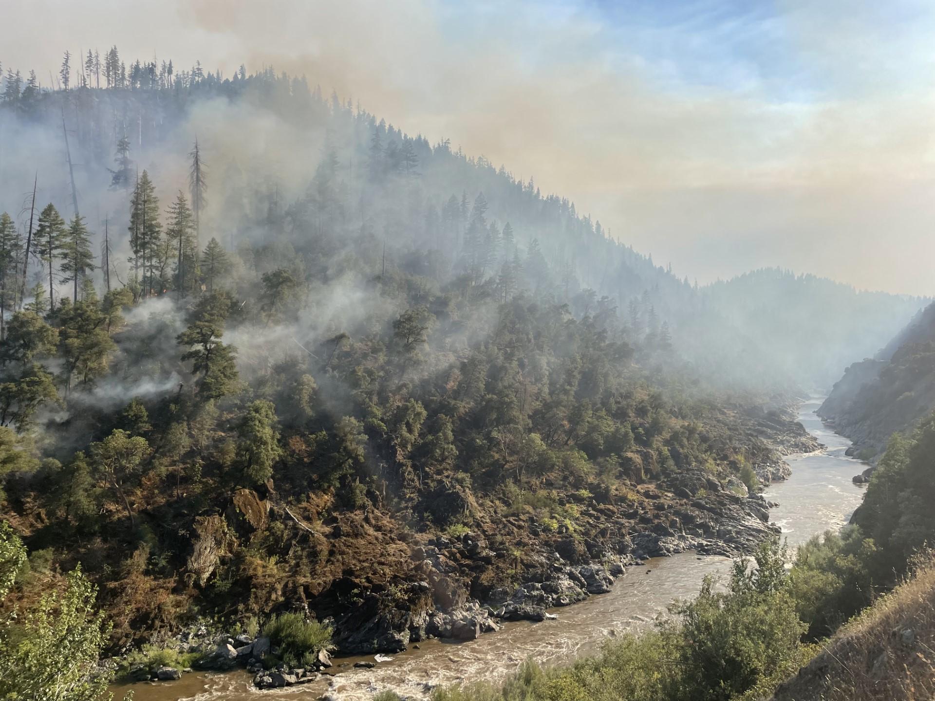 Image of the Klamath River with fire burning under the trees on the steep slopes across the river. Photo USDA Forest Service courtesy Shane McCann