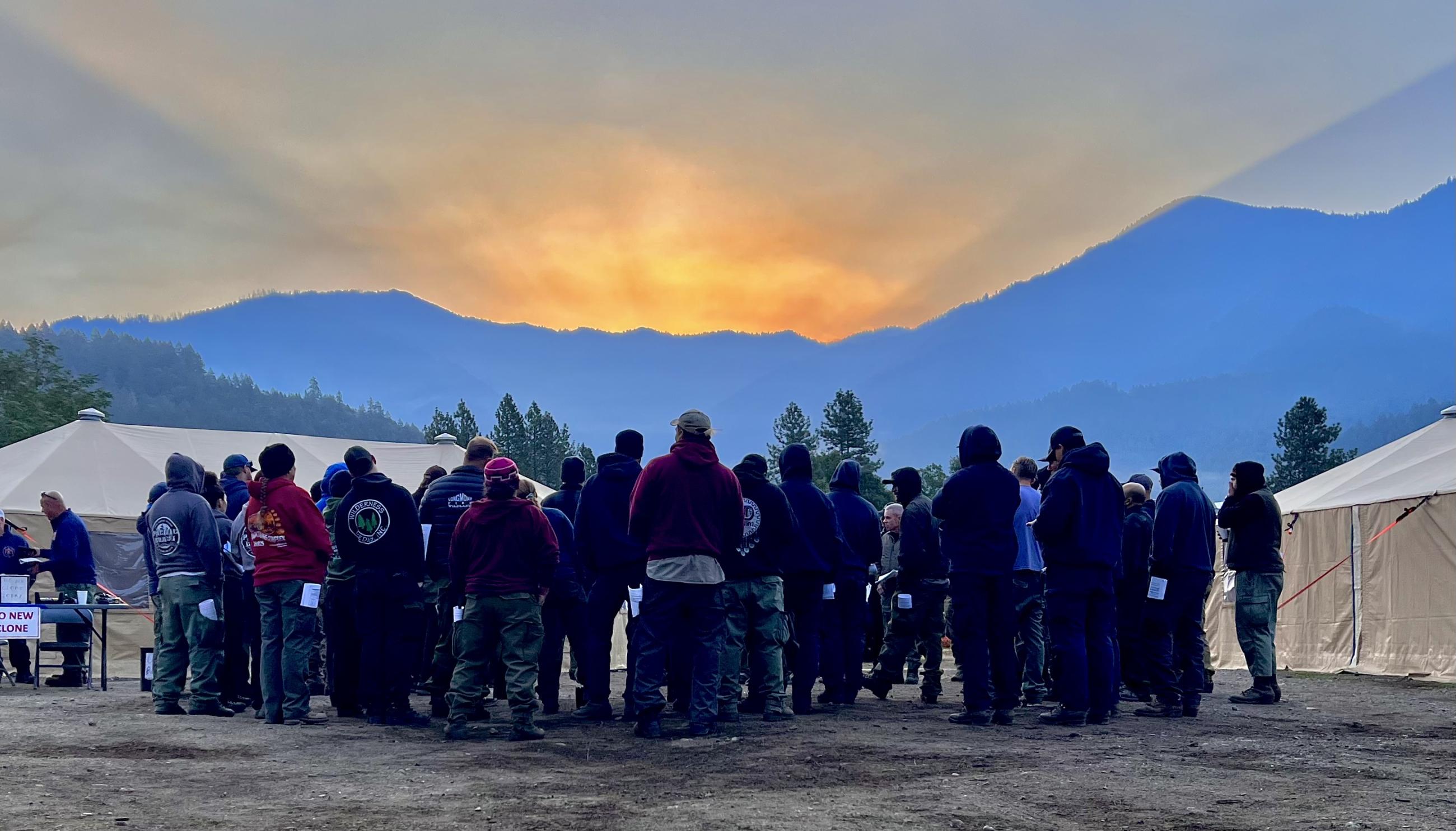 A large group of firefighters standing between two yurts as the sun rises over the mountains