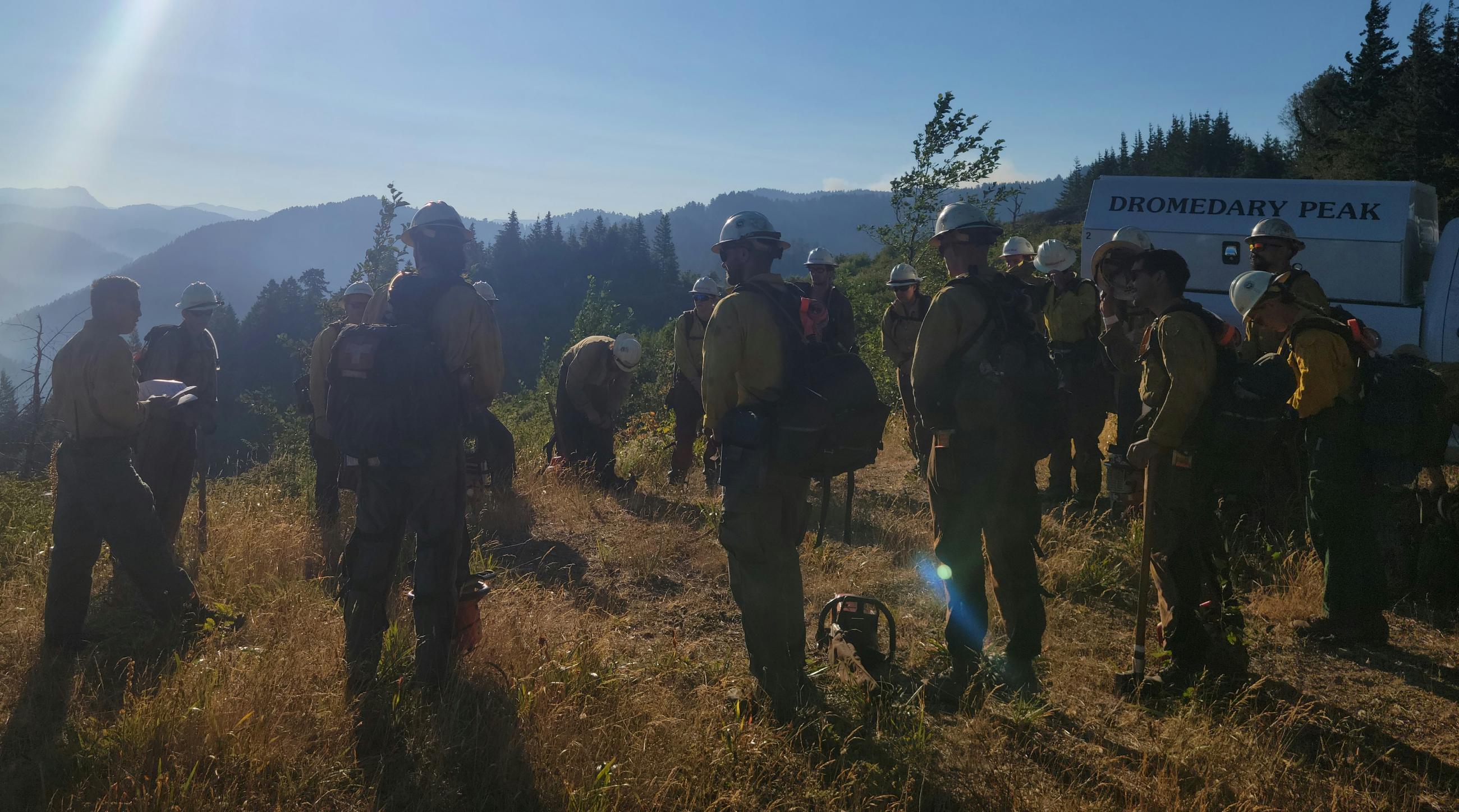17 wildland firefighters stand in a circle next to a fire vehicle with the name Dromedary Peak on the side. Firefighters are wearing green Nomex pants and yellow shirts. Some are wearing hard hats and a chainsaw is on the ground.