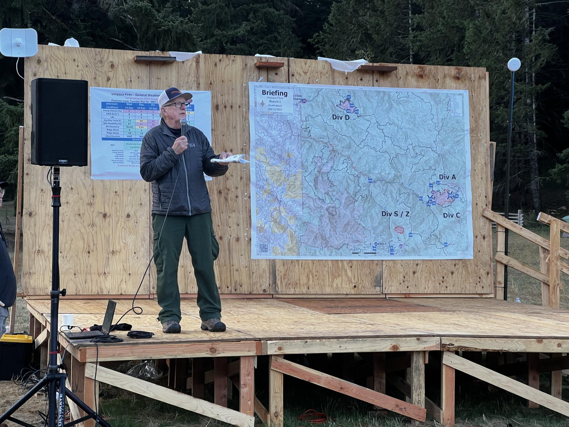 A fire manager speaks into a microphone on stage with a map posted behind him