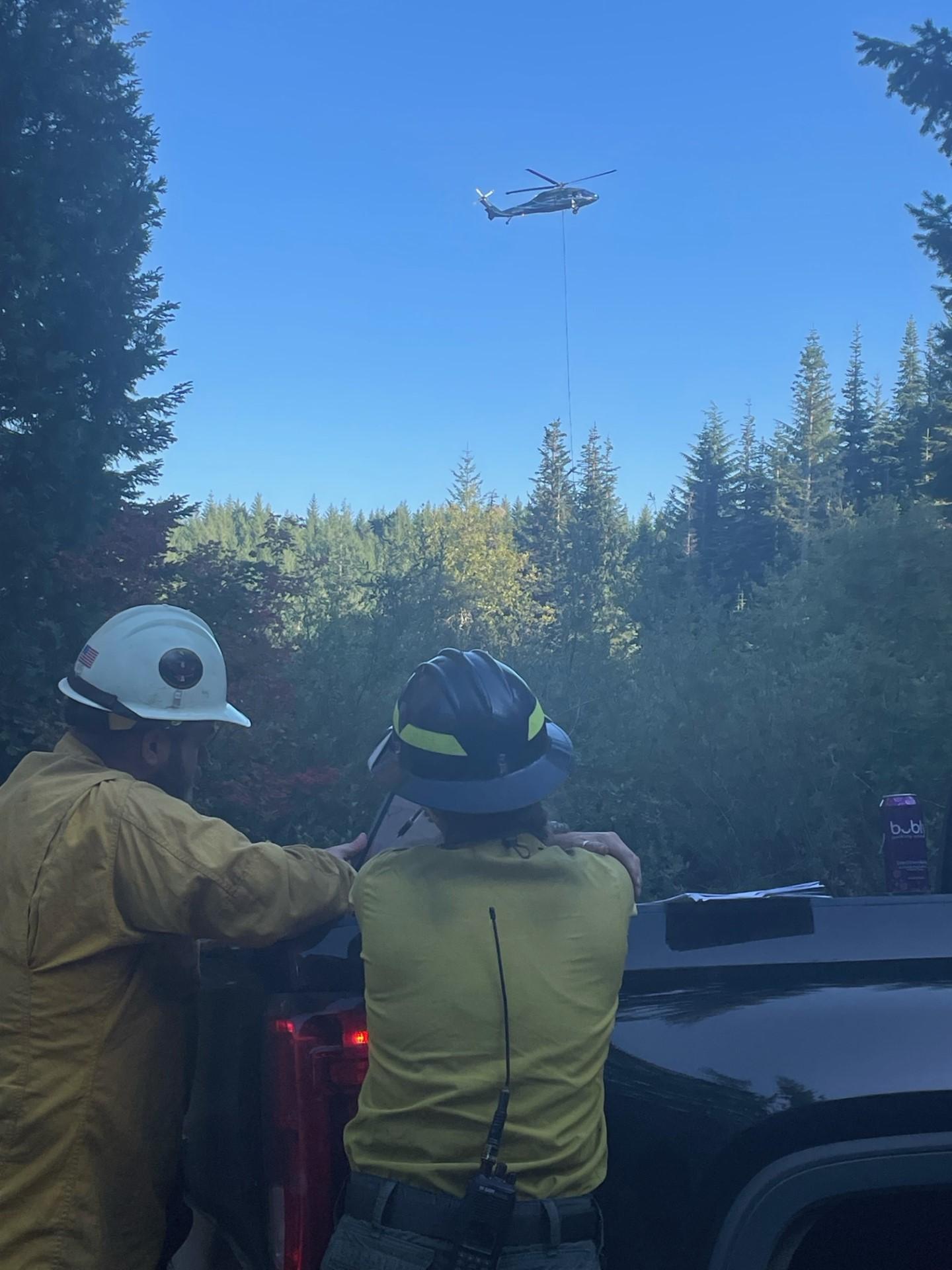 Two firefighters look at a document over a truck bed while a helicopter pulls a bucket in the distance