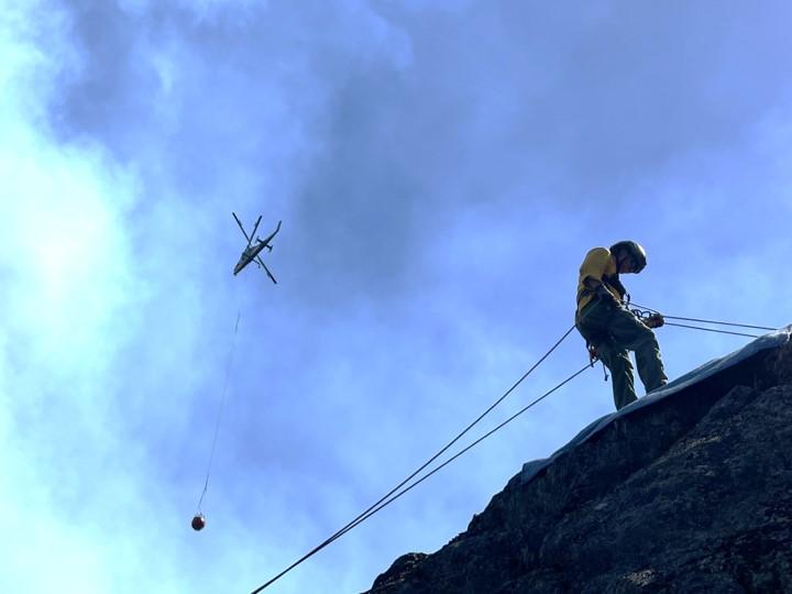A first  responder repels  down a rock face while a helicopter transports a bucket of water in the background