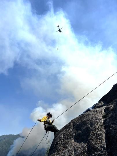 A first responder repels down a rock face  while a helicopter transports a bucket of water in the background