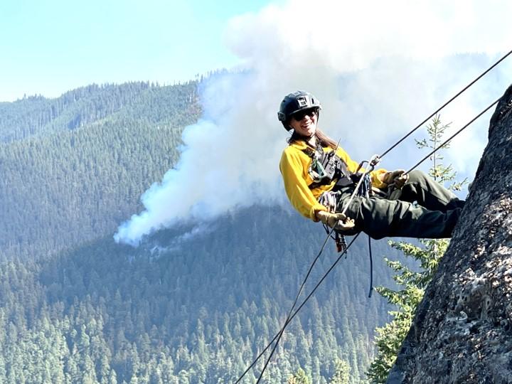 A first responder repelling down a rock face pauses to smile for the camera. Wildfire smoke rises in the background.