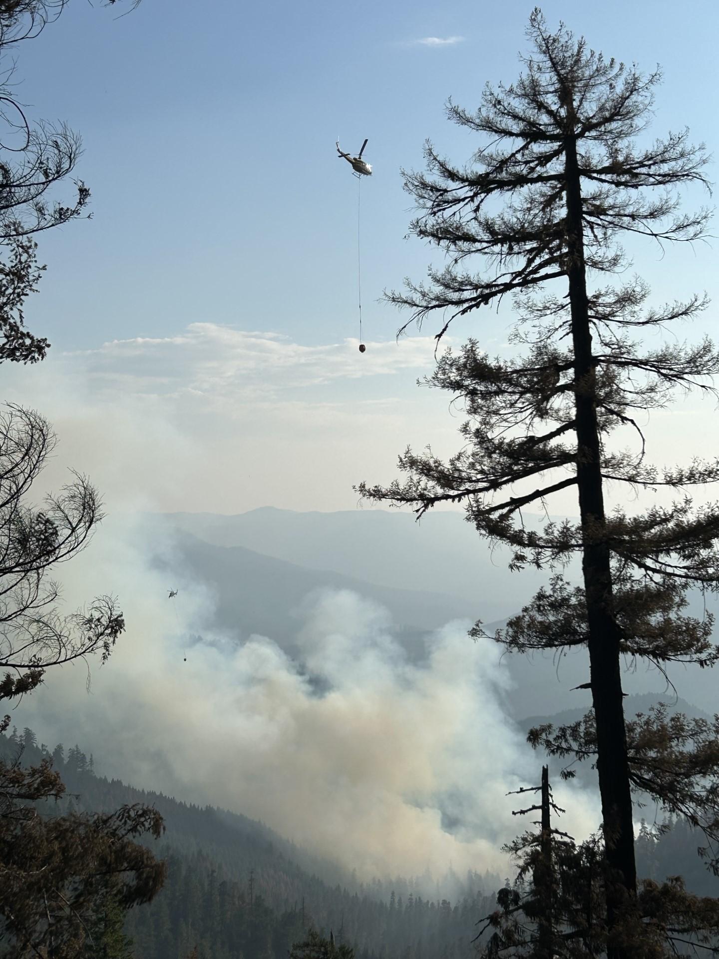 A helicopter performs bucket work over a smoky area