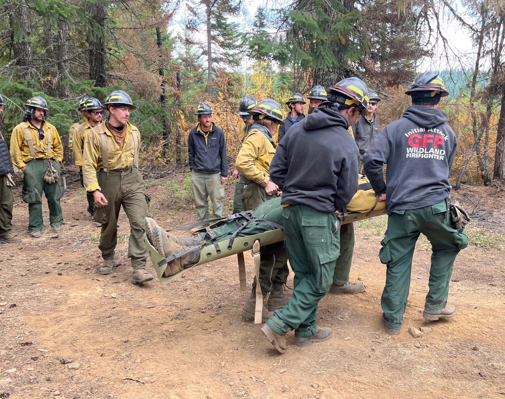 Four firefighters carry an individual on a backcountry stretcher while about a dozen other firefighters observe