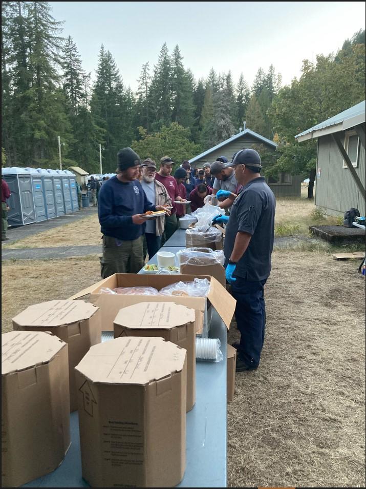 Firefighters line up to eat at Pine Creek Spike Camp. Crews are stationed there because they are working the southern fires, saving daily travel time from the main fire camp. Their meals come in "hot cans" (the round cardboard in the foreground) from off site, allowing crews to enjoy a hot meal after a long shift.