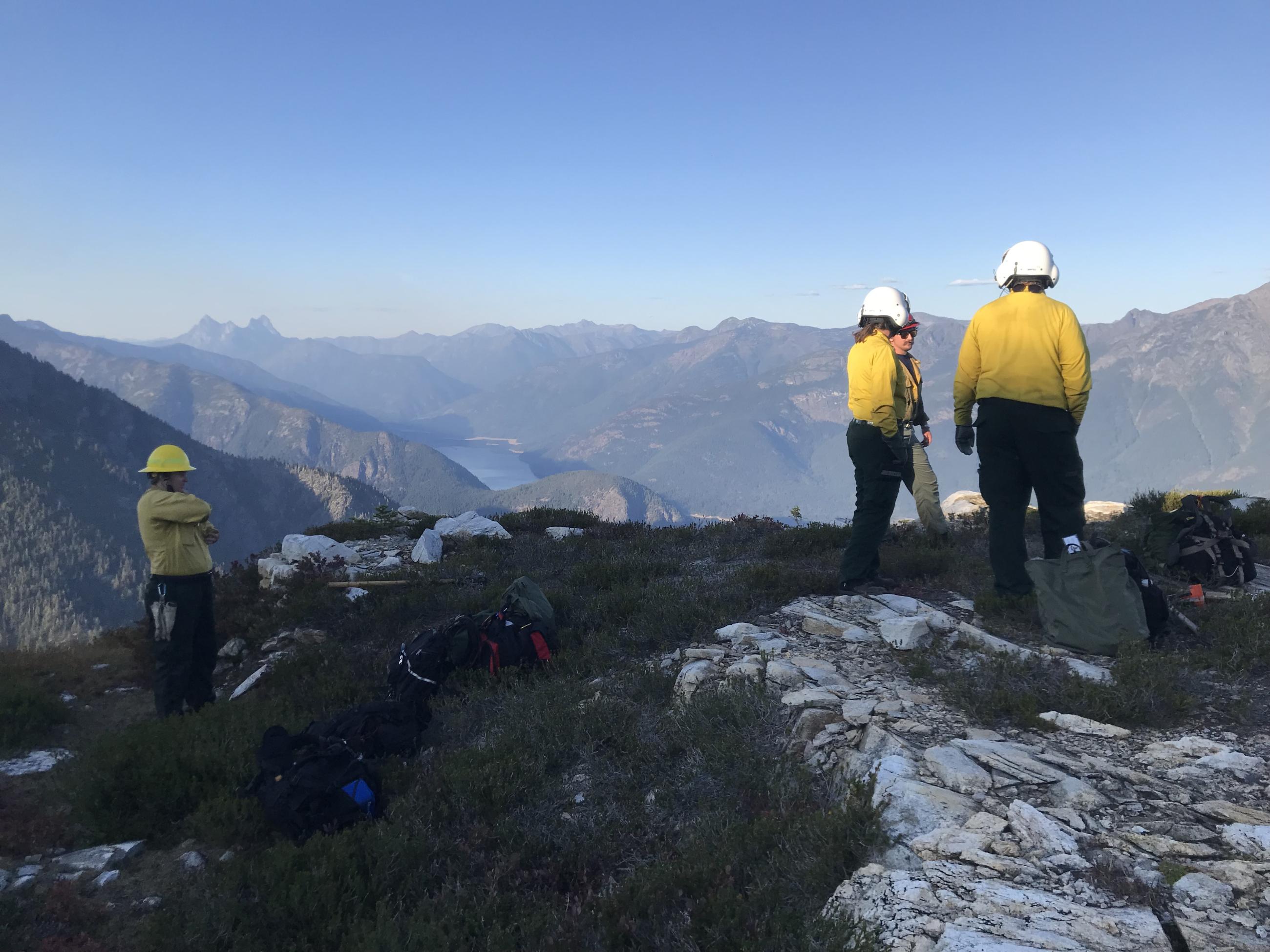 Four people in yellow shirts, green pants, and helmets stand on a rocky section of mountain with blue sky beyond them