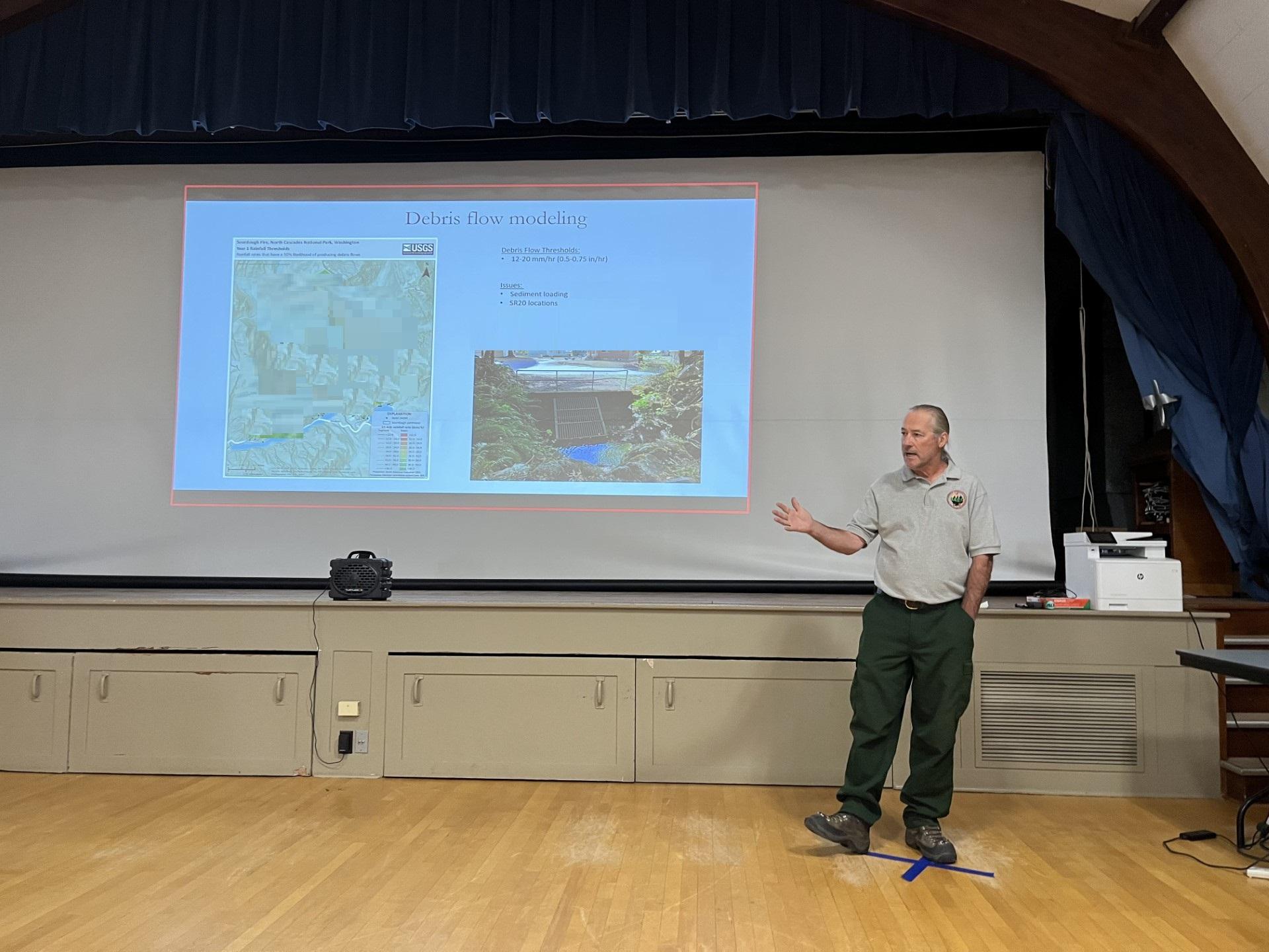 A man with green pants and a tan polo shirt stands in front of a screen with maps