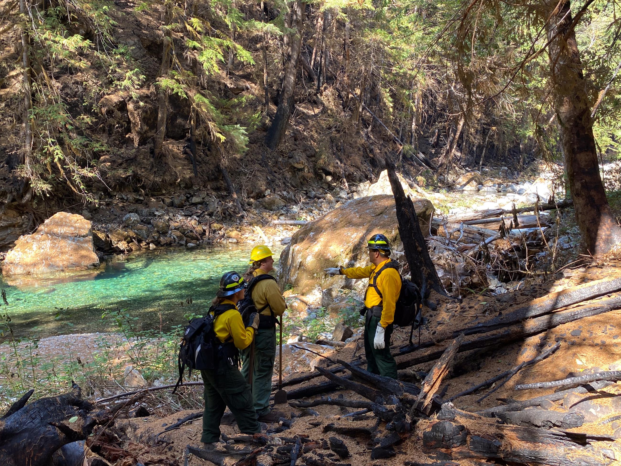 Three people in yellow shirts and helmets stand in a burnt forest next to a bright blue, clear creek