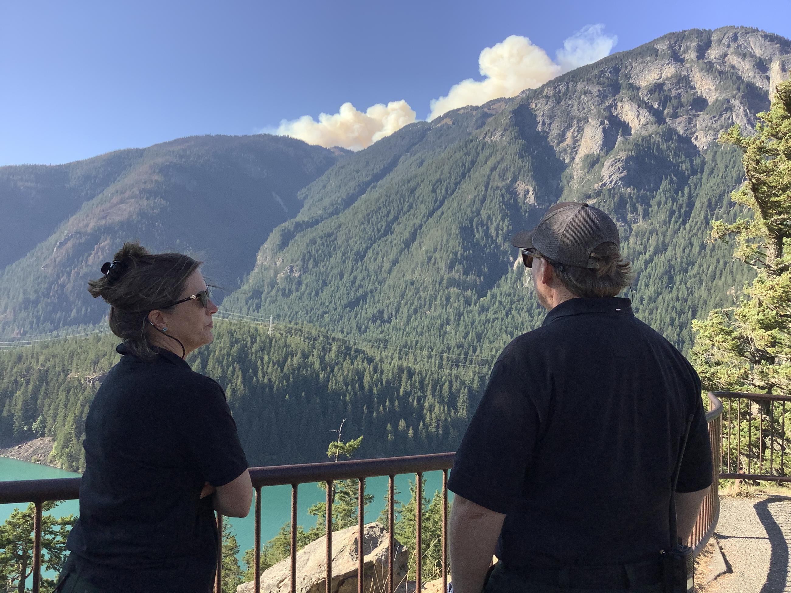 Two people in dark polo shirts stand behind a low fence, overlooking a blue body of water with wildfire smoke rising over a mountain in front of them