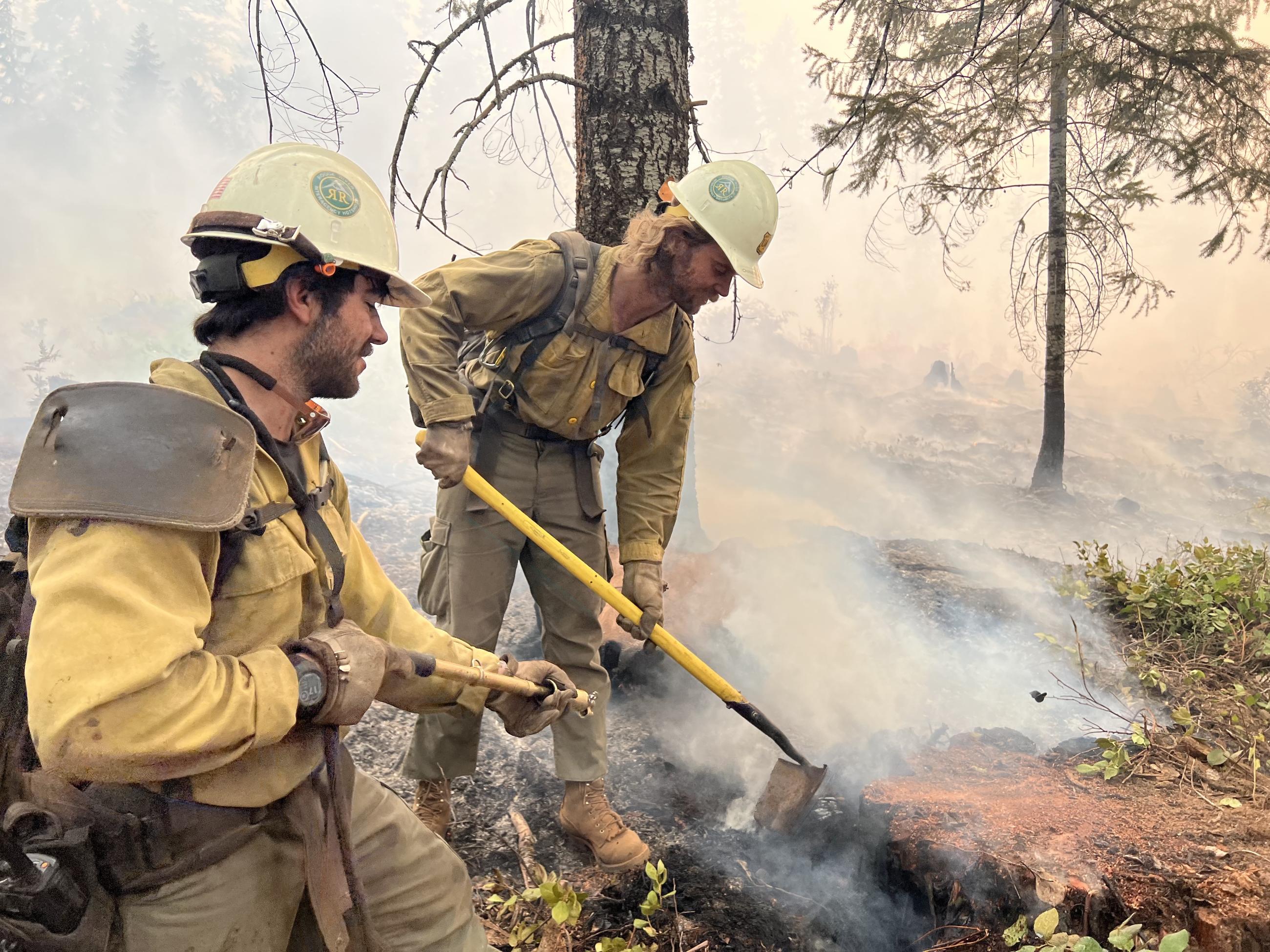 Two firefighters, one with a tool and another with a bladder bag (used to spray small amounts of water) work around a stump.