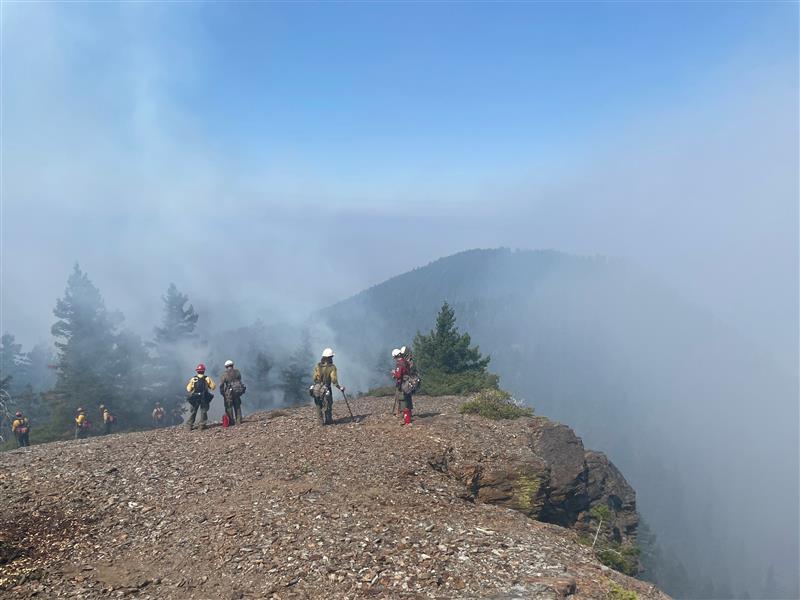 Firefighters in green and yellow clothing standing on an embankment in the mountains with smoke all around