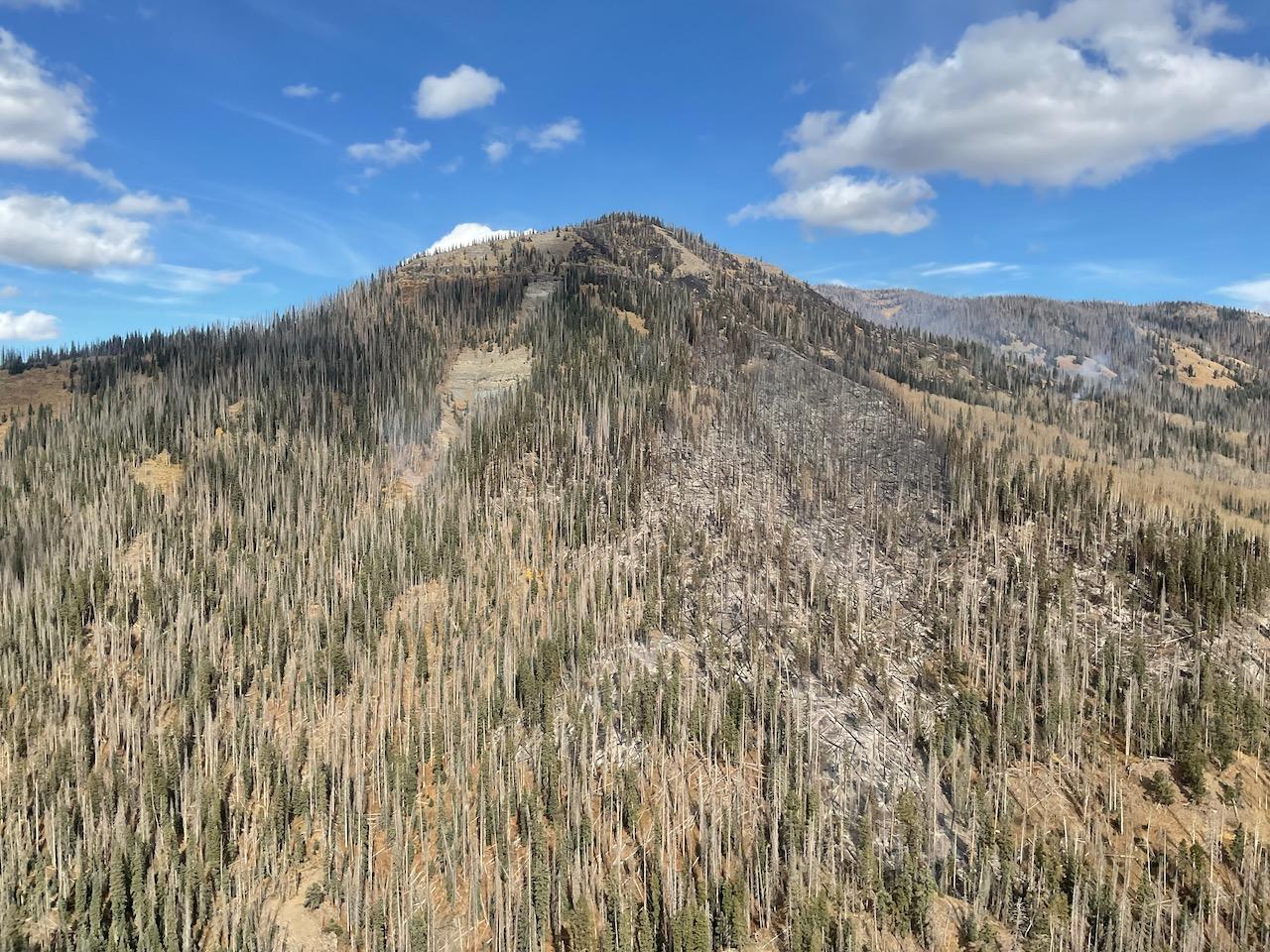 This is an aerial view showing several whisps of smoke rising from a rocky mountain peak, along with a blackened area that previously burned.