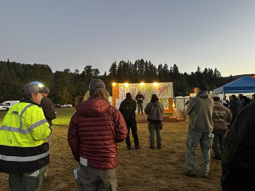 Crews stand gathered aroundd a speaker on a platform with a map behind them at a briefing