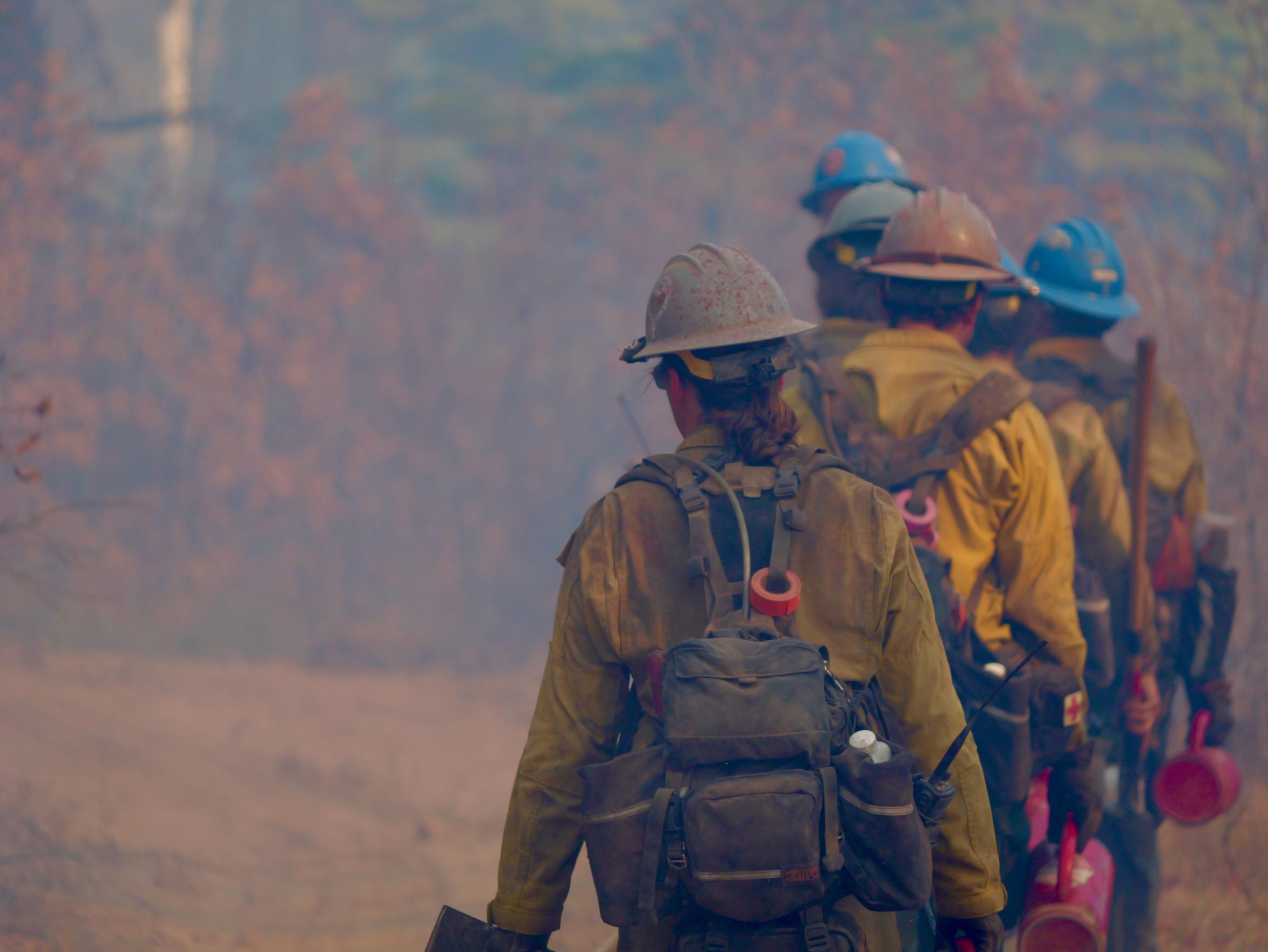 Five firefighters are seen, in grey and blue helmets and wearing packs, walking away down a road shrouded in smoke.