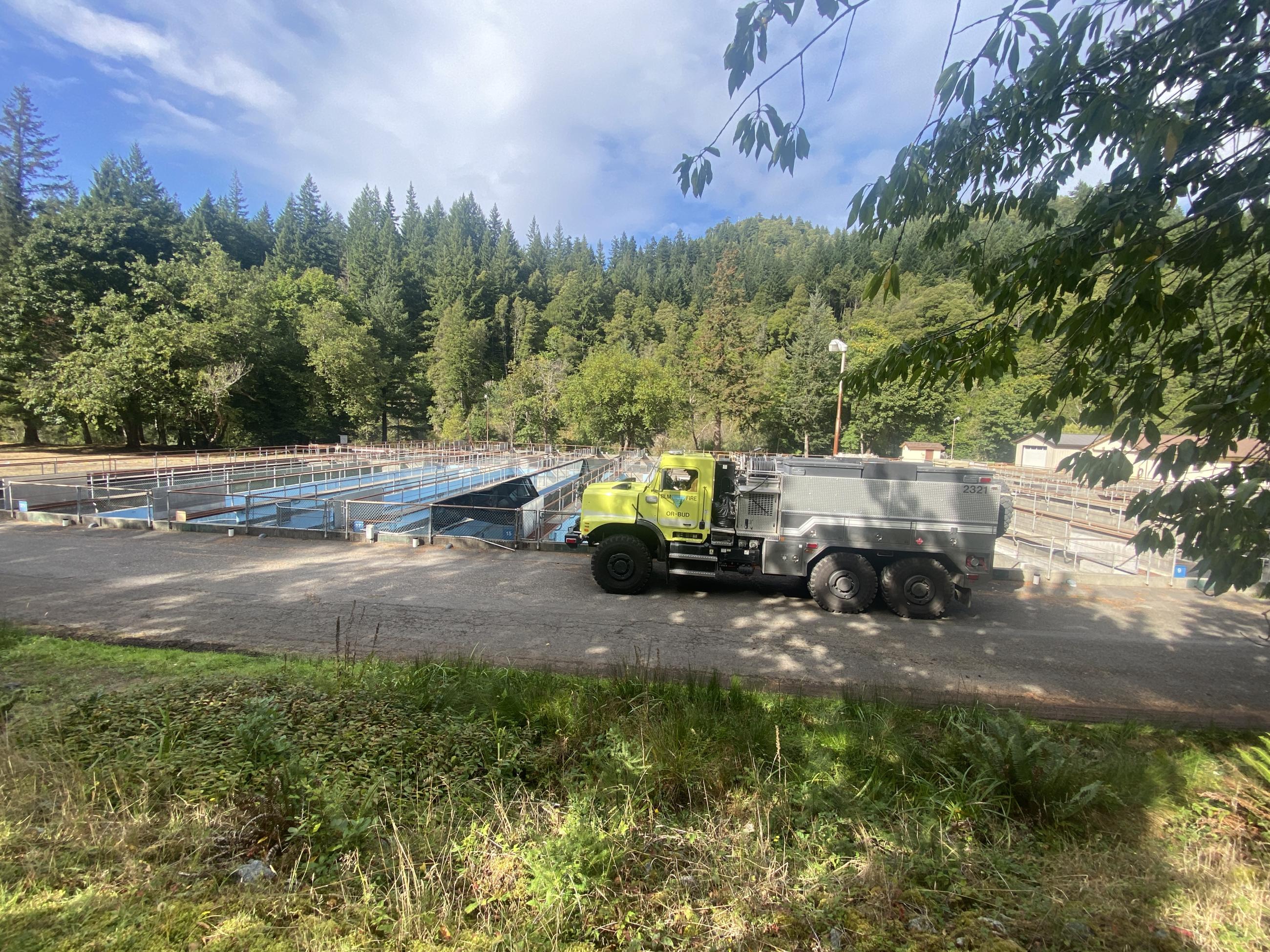 A large yellow fire engine is parked next to ground tanks at a fish hatchery