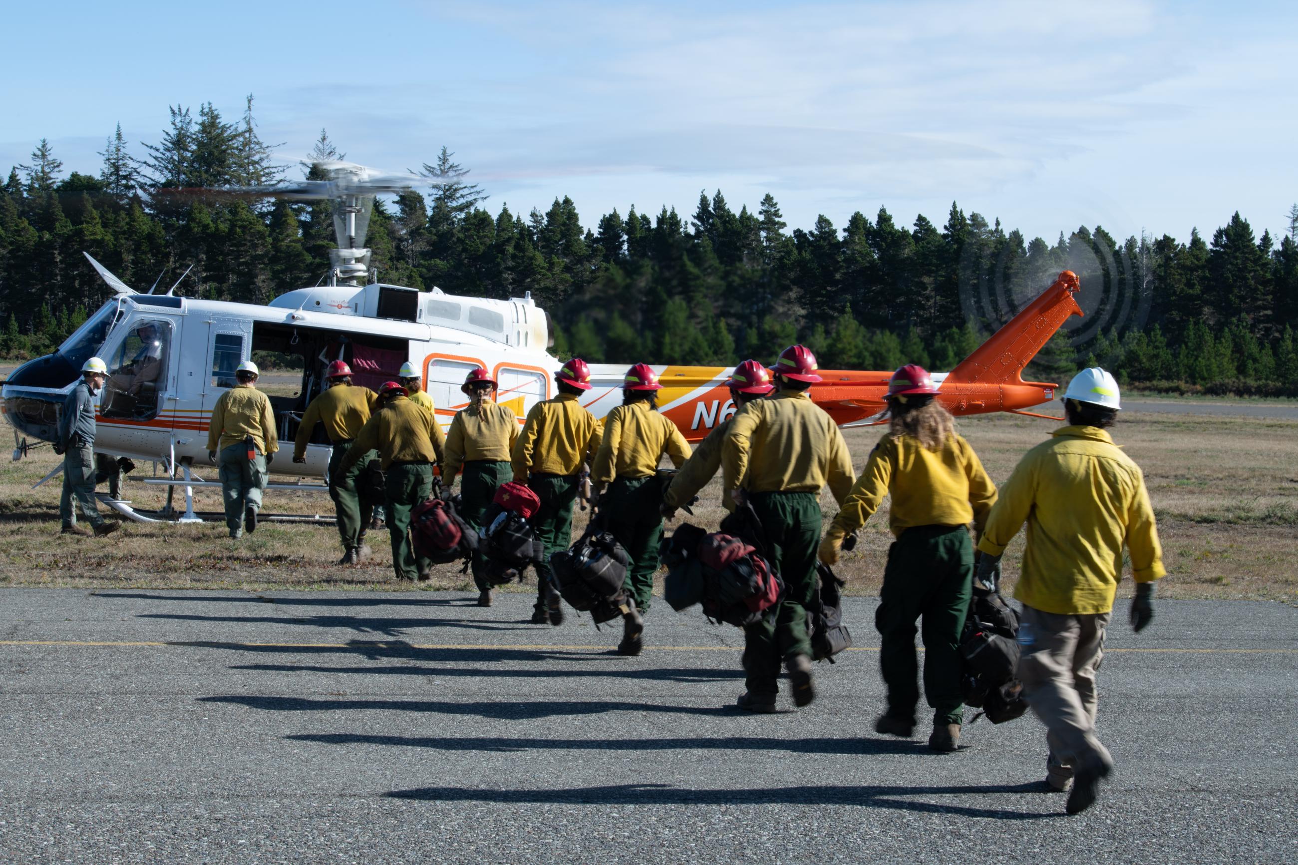 firefighters walk in a line towards a helicopter and carrying bags