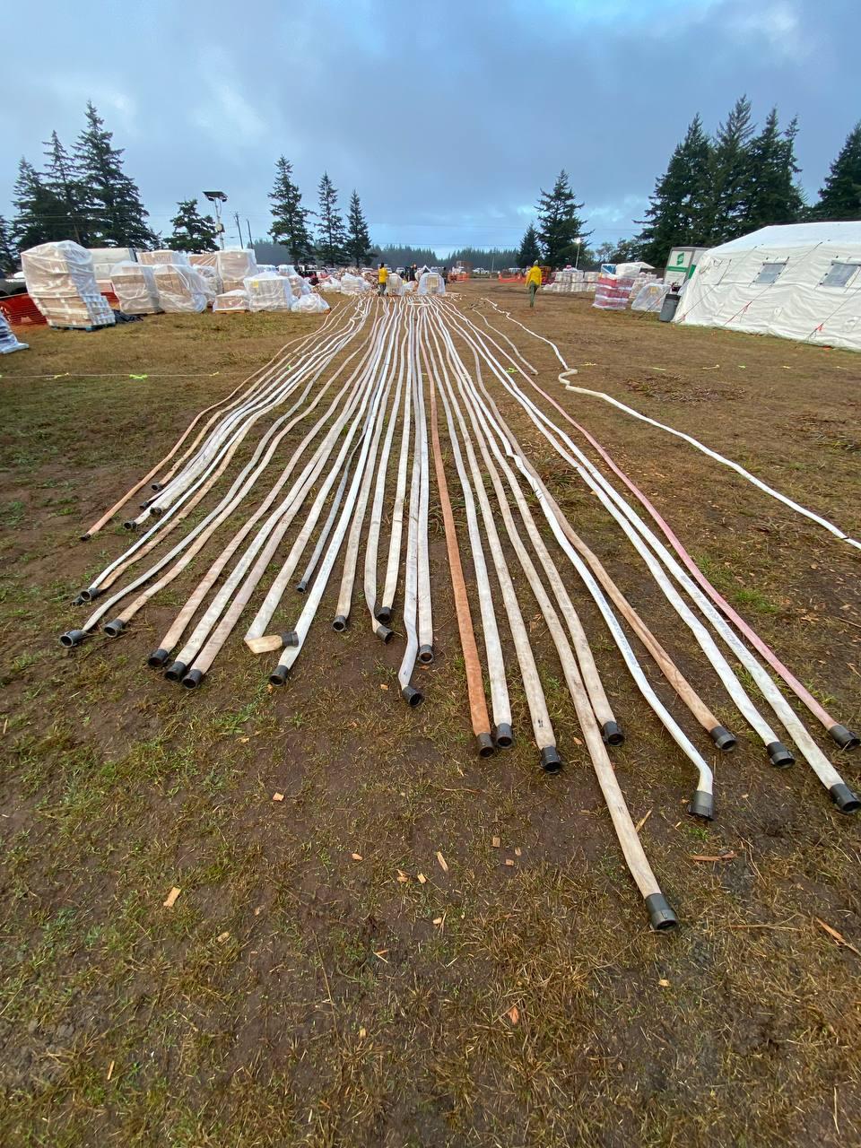 Over a dozen hoses are laid out side by side on the ground.  Stacked pallets of supplies are located to the left with a yurt to the right. Background is cloudy sky.