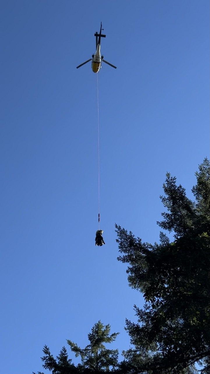 A crewmember is suspended on a line below a helicopter against a blue sky.