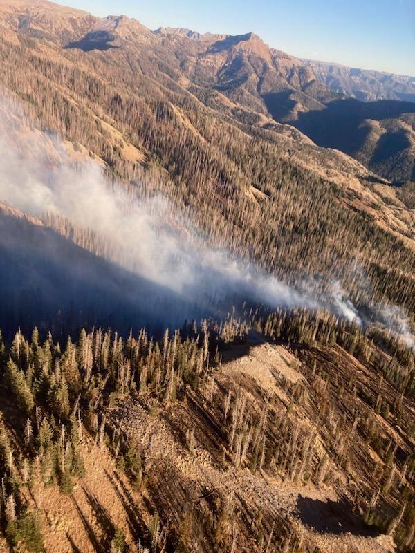 The image shows an aerial view over a mountainous terrain with smoke rising from a drainage.