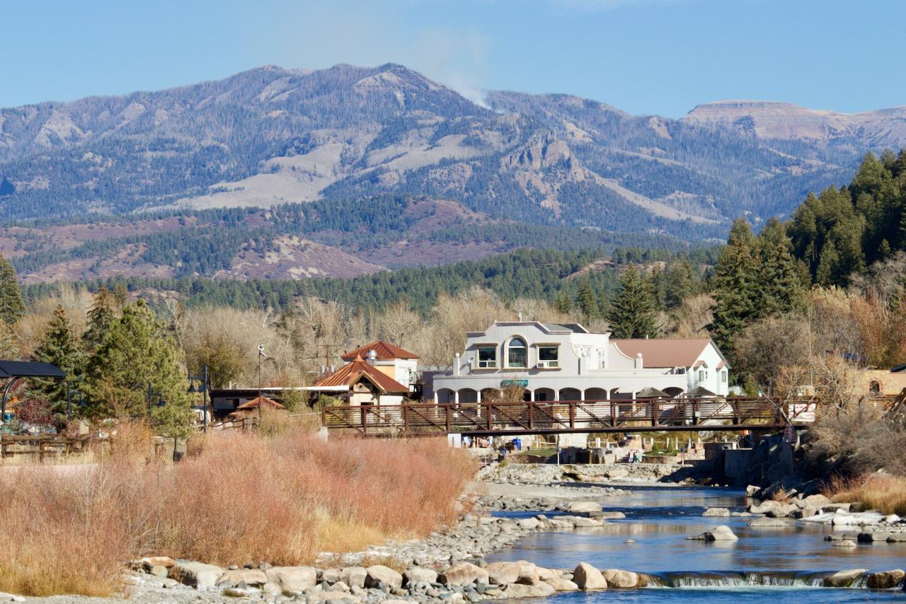 The image shows a river in the foreground with vegetated banks, a spa in the mid ground, and a mountain peak in the background with a plume of smoke rising from it.