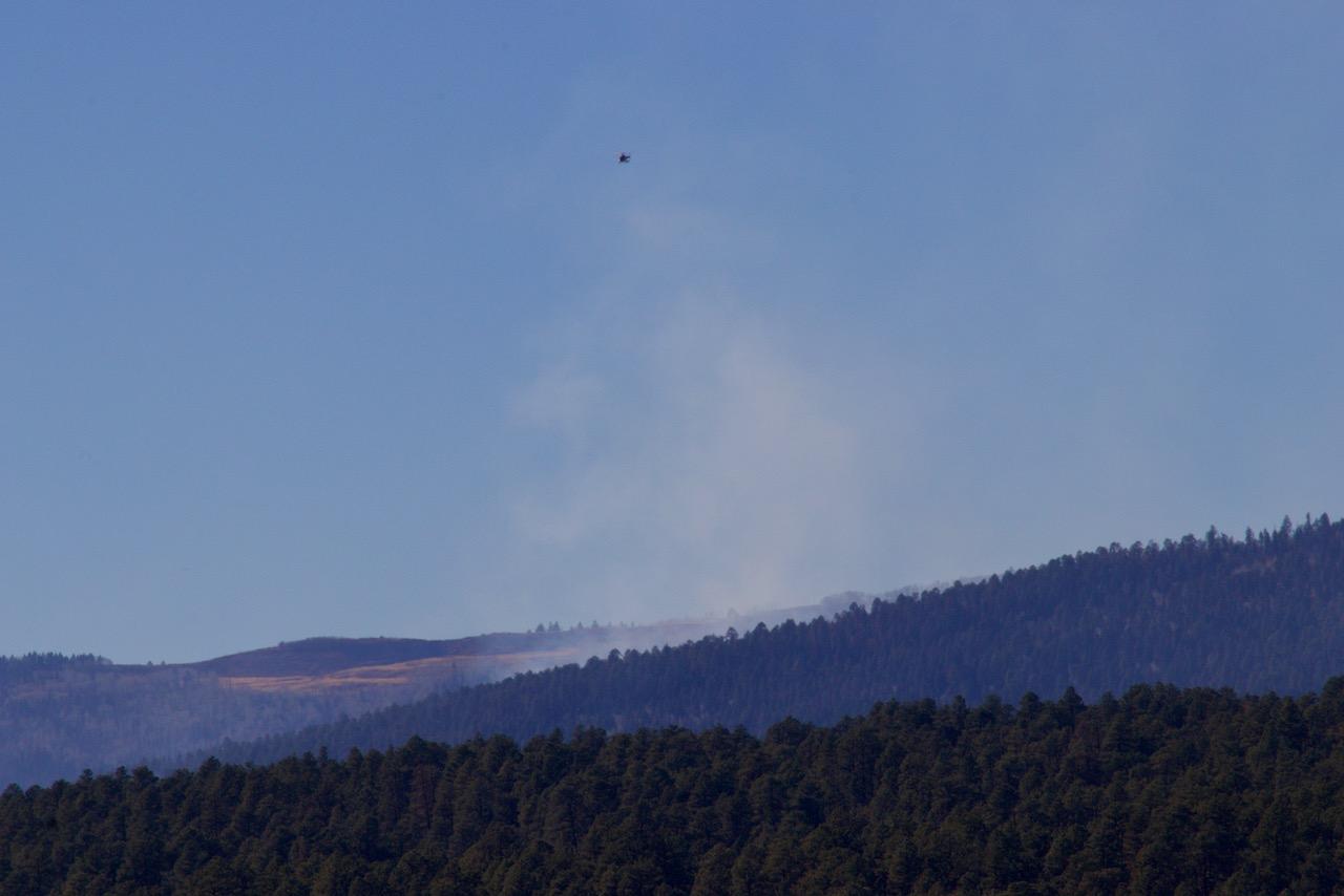 Rolling, forested hills are pictured with smoke rising from a drainage and a tiny silhouette of a helicopter above