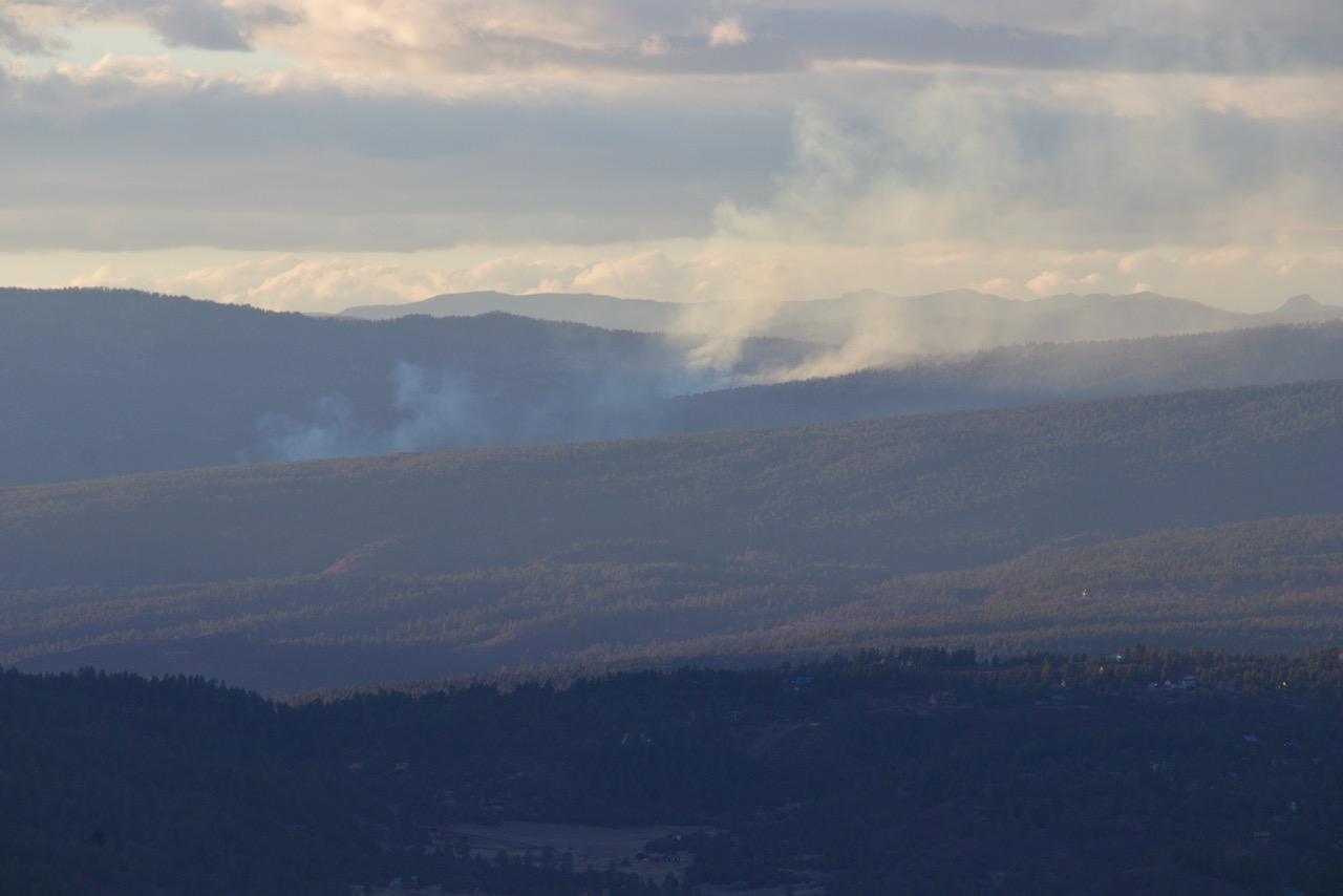 The image shows rolling, forested hills with smoke plumes rising from a drainage and an overcast sky in late afternoon light.
