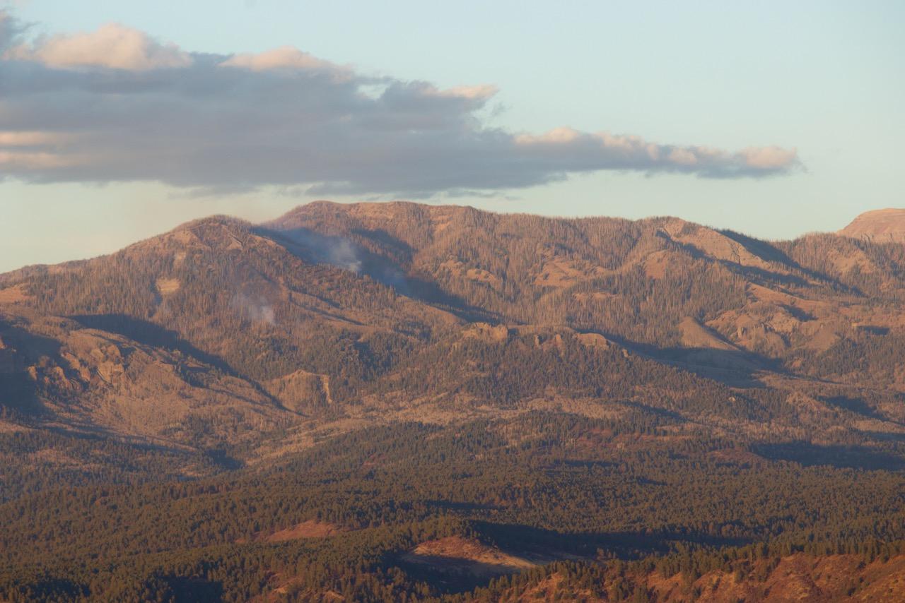 The image shows mountanous, forested terrain with craggy peaks and pastures in the valley. Whisps of smoke rise from the sides of the peak.