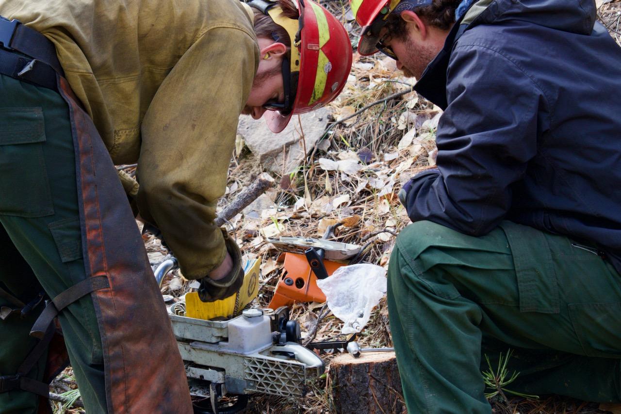 Two young men wearing green pants, yellow shirts, and red hard hats (one with orange chaps on and one with a blue rain jacket on) are crouched over a chainsaw with the dust cap removed.