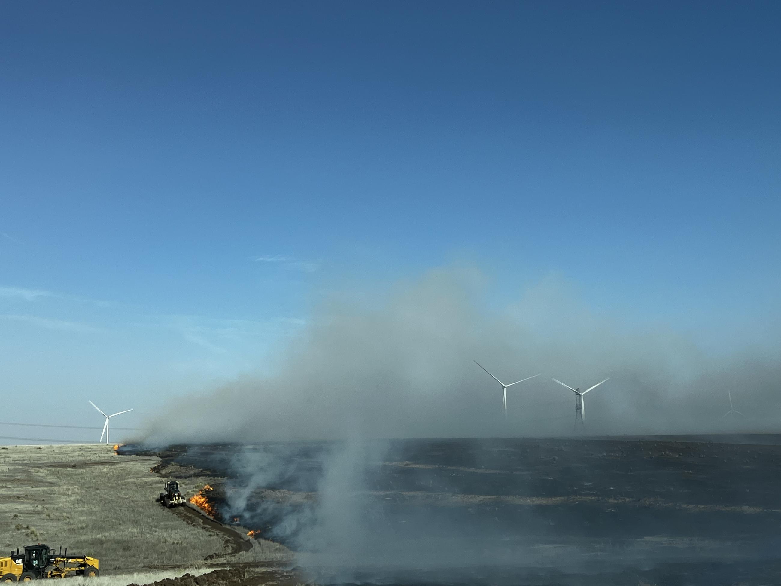 Image depicts a large field that is halfway burned, with smoke rising from the burned side. 