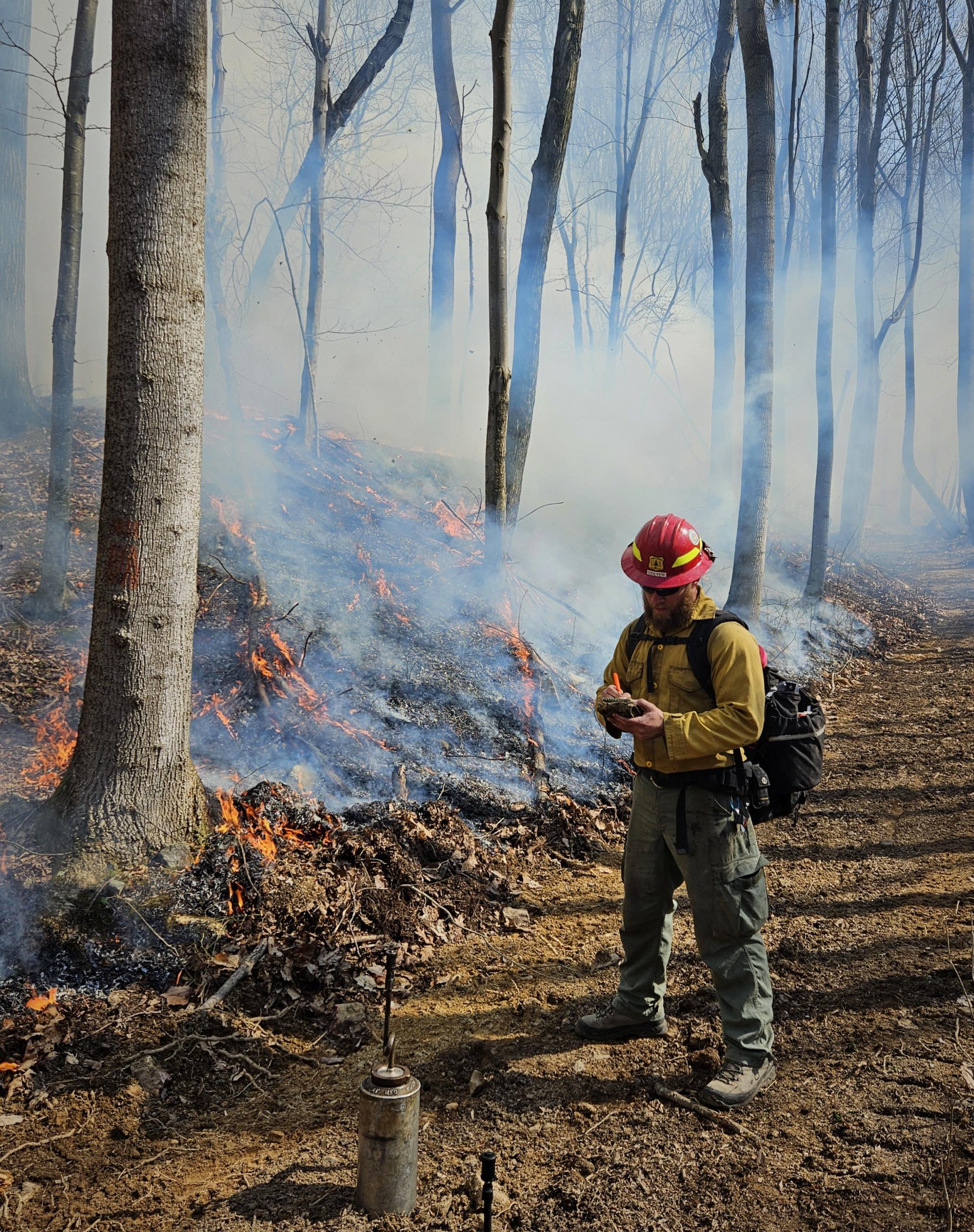 Firefighter writing in notebook standing on trail next to forested slope with short flames and smoky background with drip torch on trail in foreground