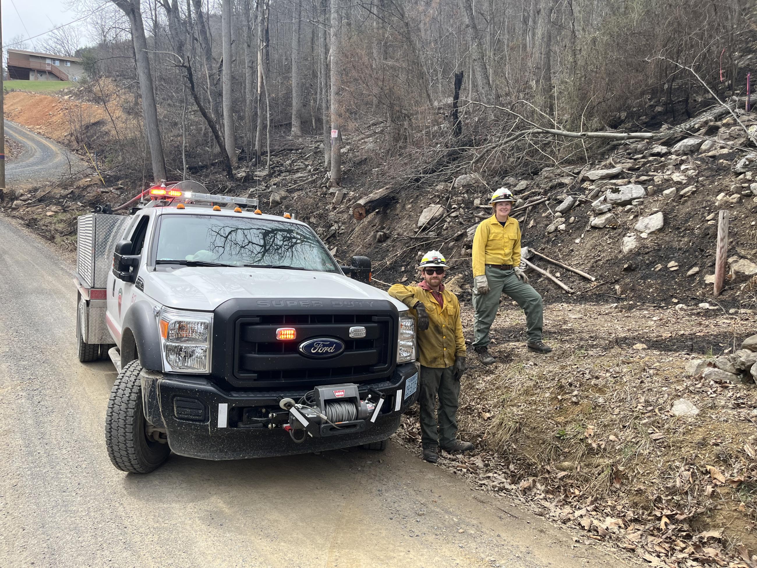 Two firefighters stand on forested slope next to truck parked down the road from a house