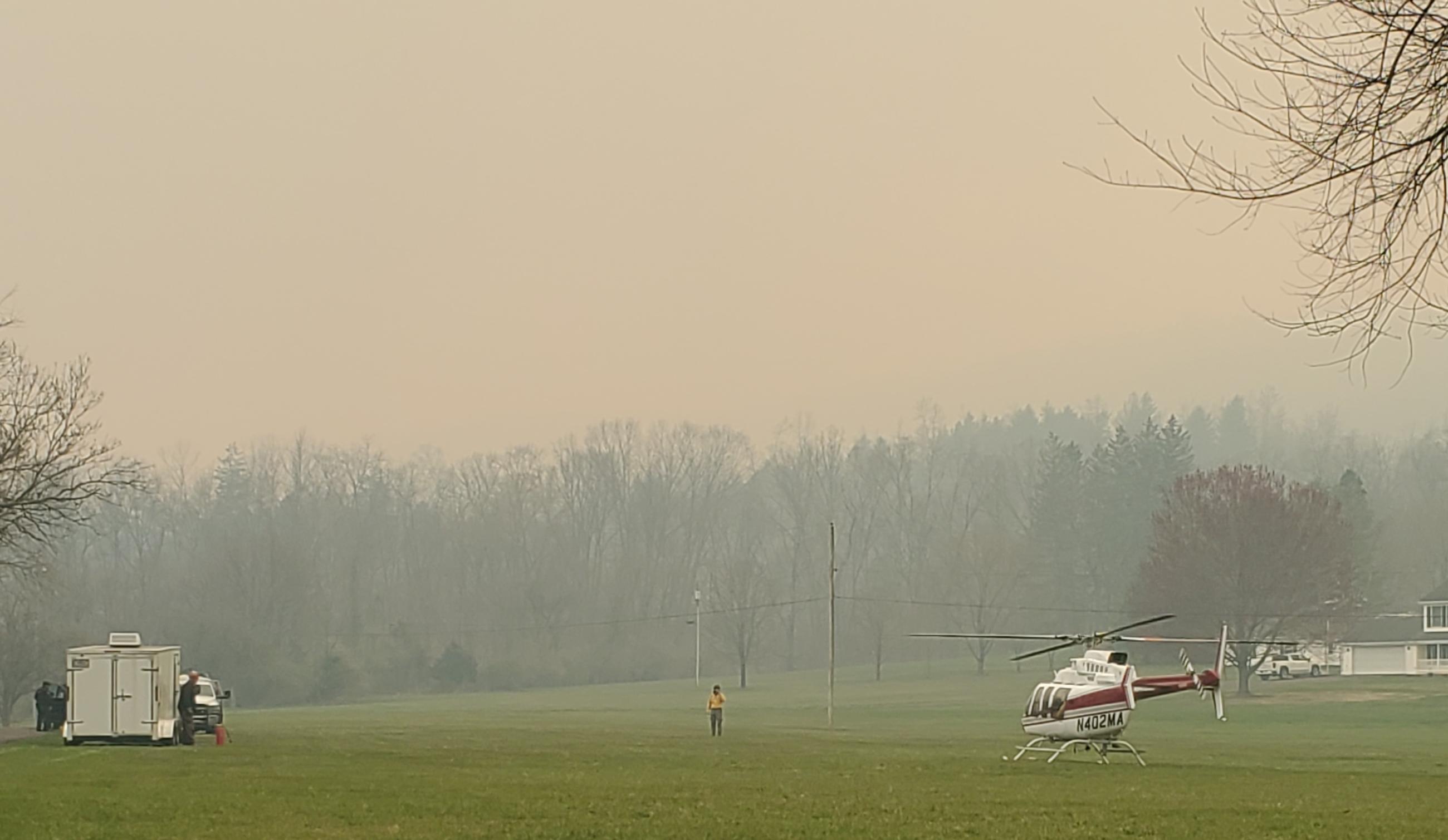 Helicopter and support vehicles on green grass under smoky sky