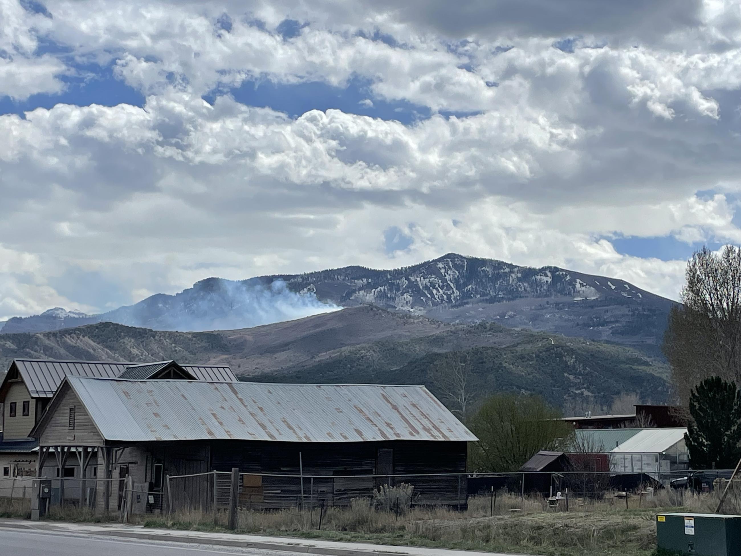 Photo shows a historic building, wood with metal roof, with mountains in the background and smoke emerging from the mountains