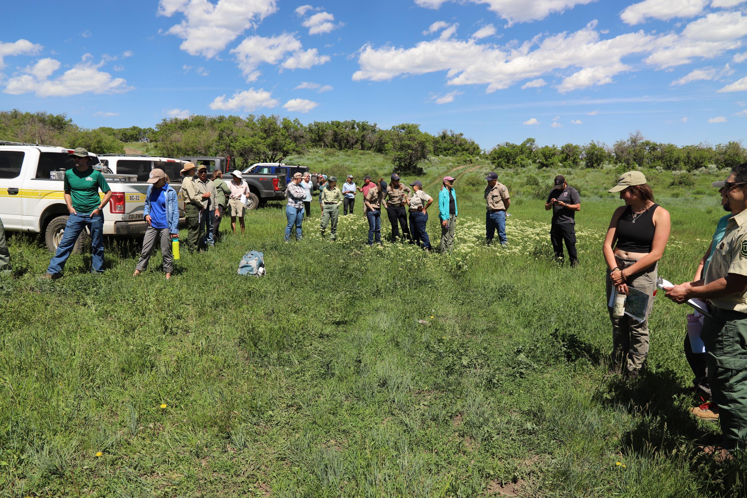 The image shows about 15 individuals standing in a grassy field watching someone speak, with a pickup truck on the left
