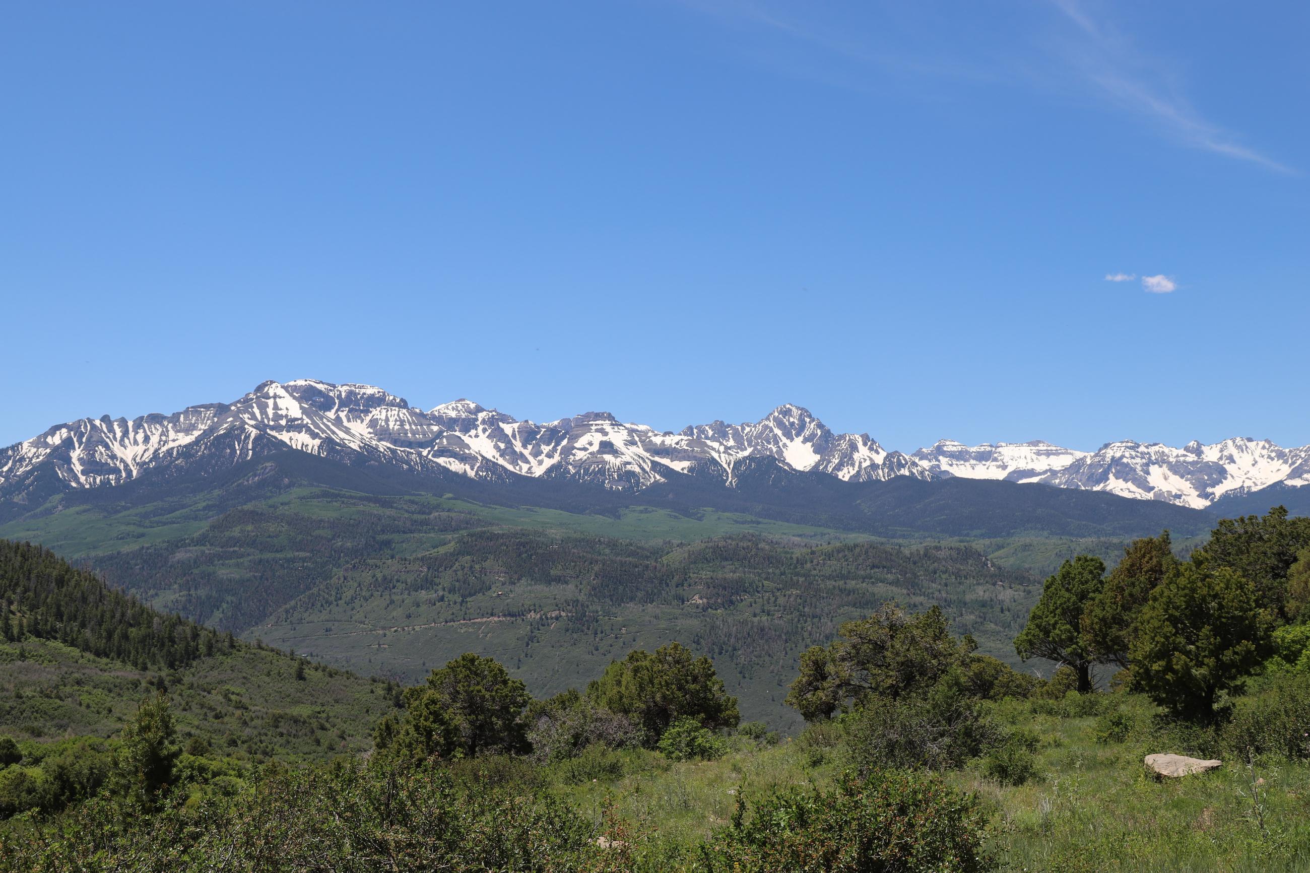 The image shows a landscape picture including ragged, snow-covered mountains in the background, green rolling hills in the foreground, and Baldy Mountain in the midground with a bluebird sky overhead