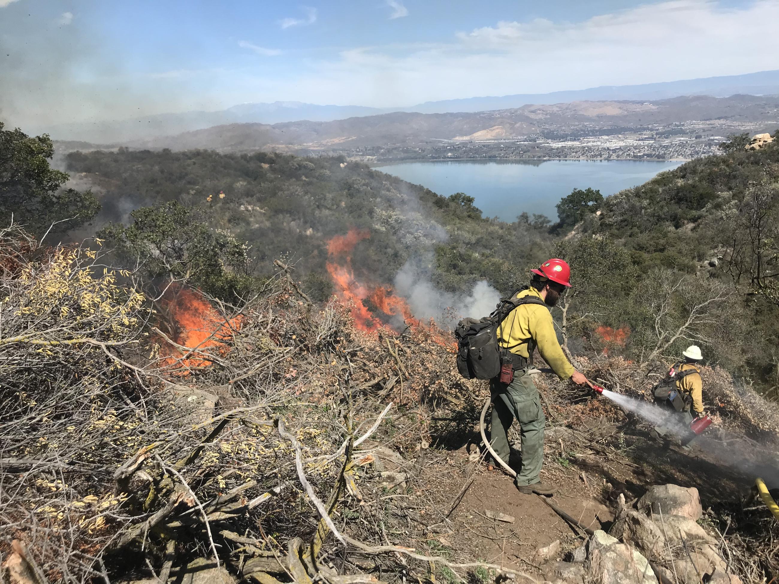 Picture shows top of a mountain with a lake in the background. There is a fighters with a hose and water coming out of it. There is fire and smoke in the background. 